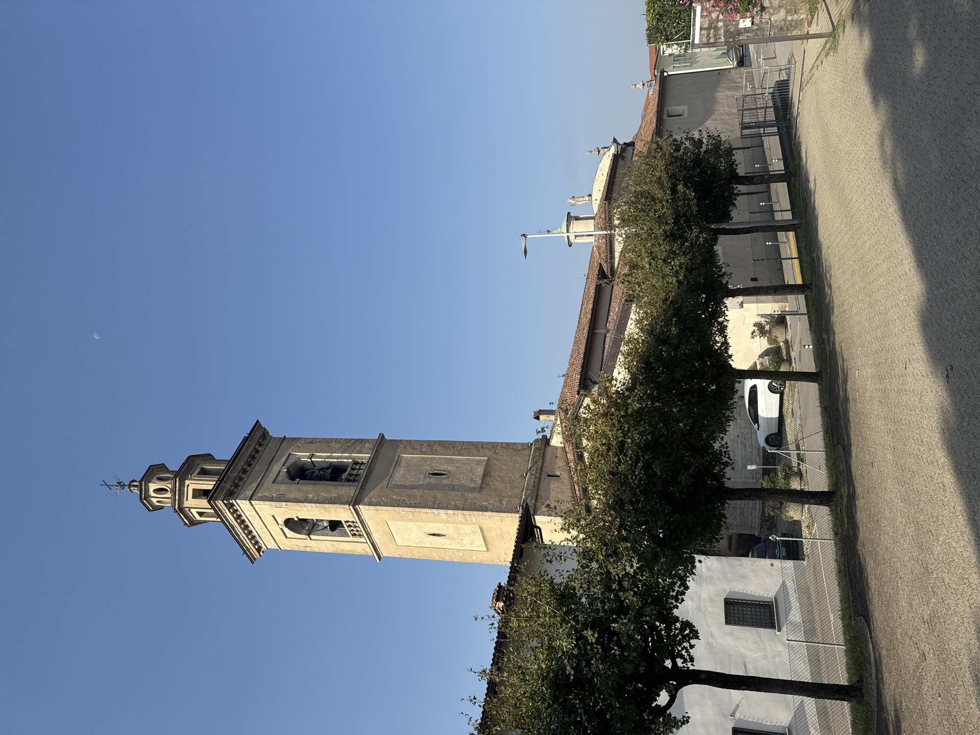 The image depicts a view of the Church of San Giorgio in Balerna, Switzerland, under a clear blue sky. A prominent bell tower, constructed of light-colored stone, dominates the left side of the frame, featuring arched openings containing bells and a decorative top with a cross. The church building itself, with a red-tiled roof, extends from the base of the tower. A line of mature trees with dark green foliage runs along the front of the church and adjacent buildings. To the left, a white building with dark-grated windows is visible. In front of the church and buildings, a paved walkway or courtyard is present, bordered by a fence. Several vehicles, including a white hatchback and a dark-colored car, are parked in the vicinity. The overall scene is tranquil and shows architectural elements of a European town.