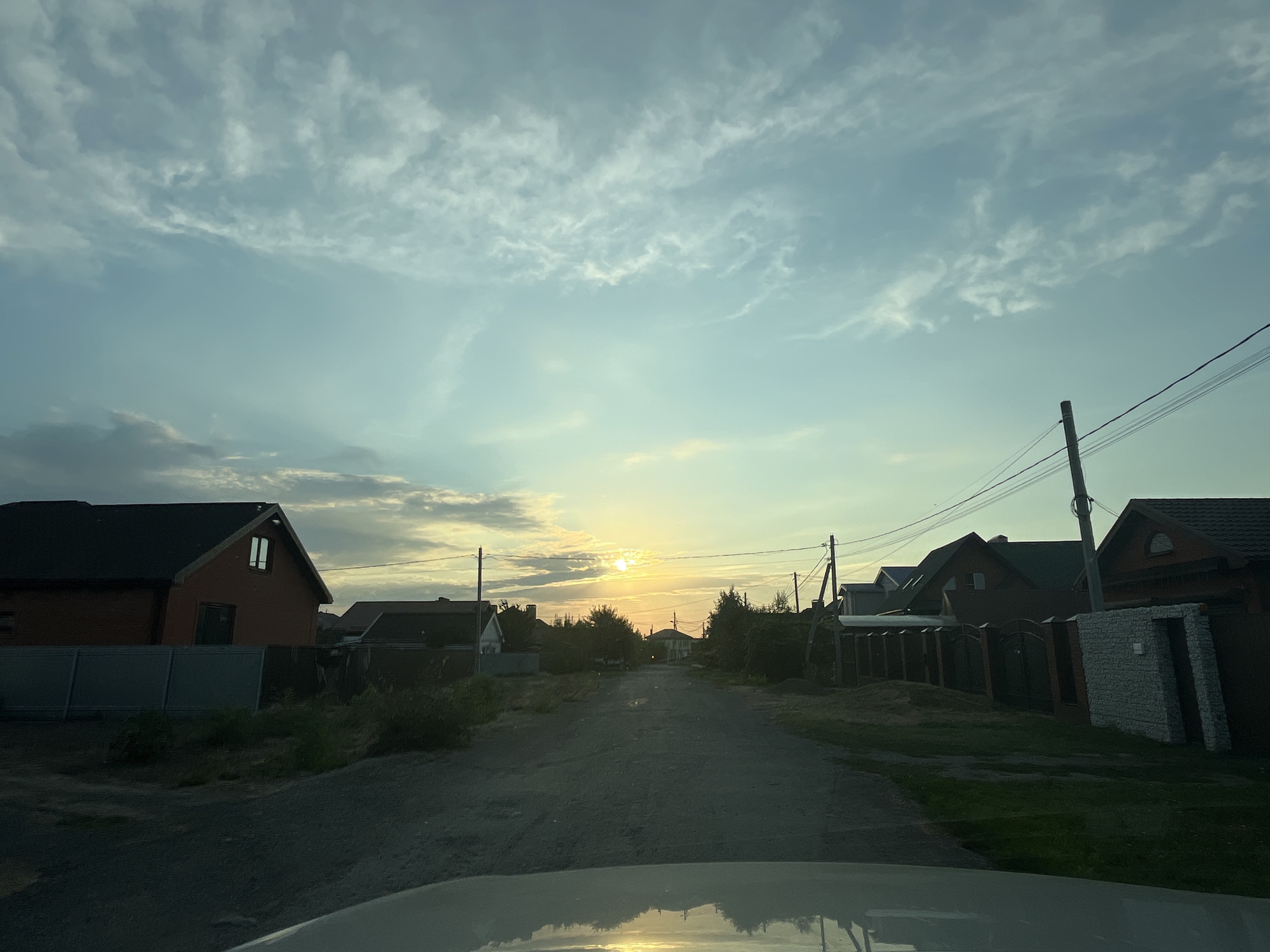 The image shows a dirt road in a residential area, likely in the city of Taganrog City District, Russia. The sun is low in the sky, creating a warm, golden light that reflects off the hood of the vehicle from which the photo is taken. On either side of the road are houses with dark-colored roofs and brick exteriors, some with decorative fences. Trees and vegetation are visible, adding to the rural feel of the scene. Power lines stretch across the sky, supported by wooden poles. The sky is a soft blue with wispy clouds, characteristic of a late afternoon or early evening. The overall impression is one of a quiet, peaceful, and somewhat undeveloped suburban or rural setting during sunset.
