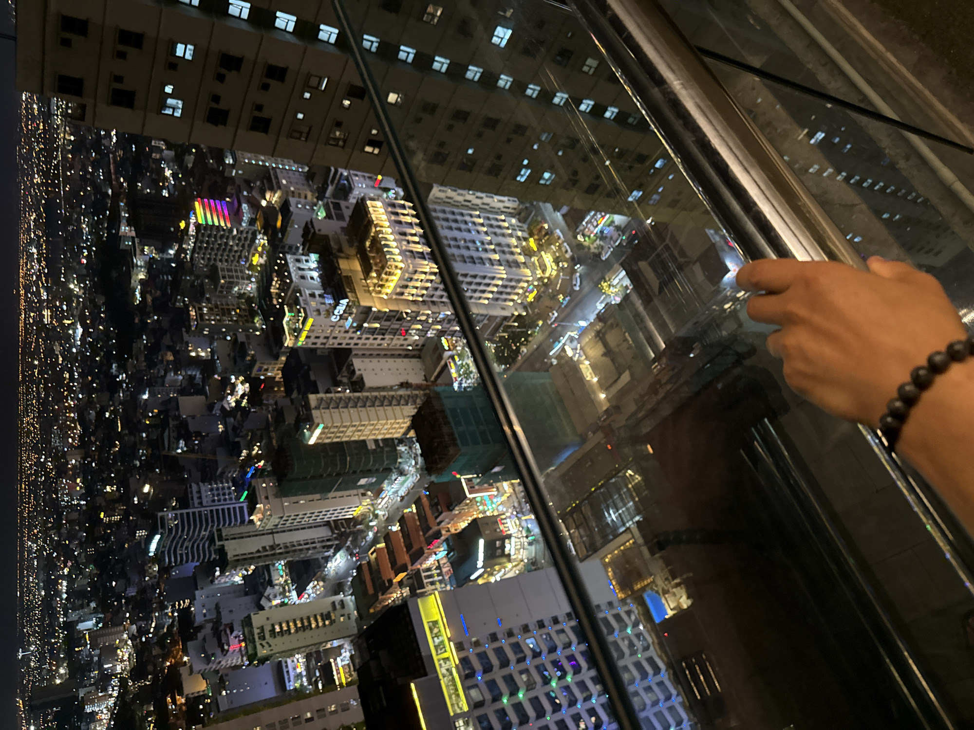 The image is a night-time aerial view of a dense urban cityscape, likely from a high vantage point such as a skyscraper or observation deck. The city is illuminated by a vast expanse of building lights, streetlights, and car headlights, creating a glittering tapestry against the dark sky.
In the foreground, a hand with a dark beaded bracelet is visible, resting on a reflective surface that appears to be glass, possibly a balcony railing or window. The hand is partially obscuring the view below.
The city below is a complex network of buildings of varying heights and architectural styles. Some buildings are modern high-rises with illuminated windows, while others appear to be older, lower structures. A particularly striking building in the mid-ground is lit with a vibrant rainbow of colors. The streets are visible, with indications of traffic. Reflections of the city lights are visible on the surfaces in the foreground, adding to the depth and visual complexity of the scene.
The overall impression is one of a bustling, vibrant city at night. Given the specific mention of Đà Nẵng, Vietnam, the scene likely depicts a view of its central business district or a similar area known for its modern development and vibrant nightlife.
