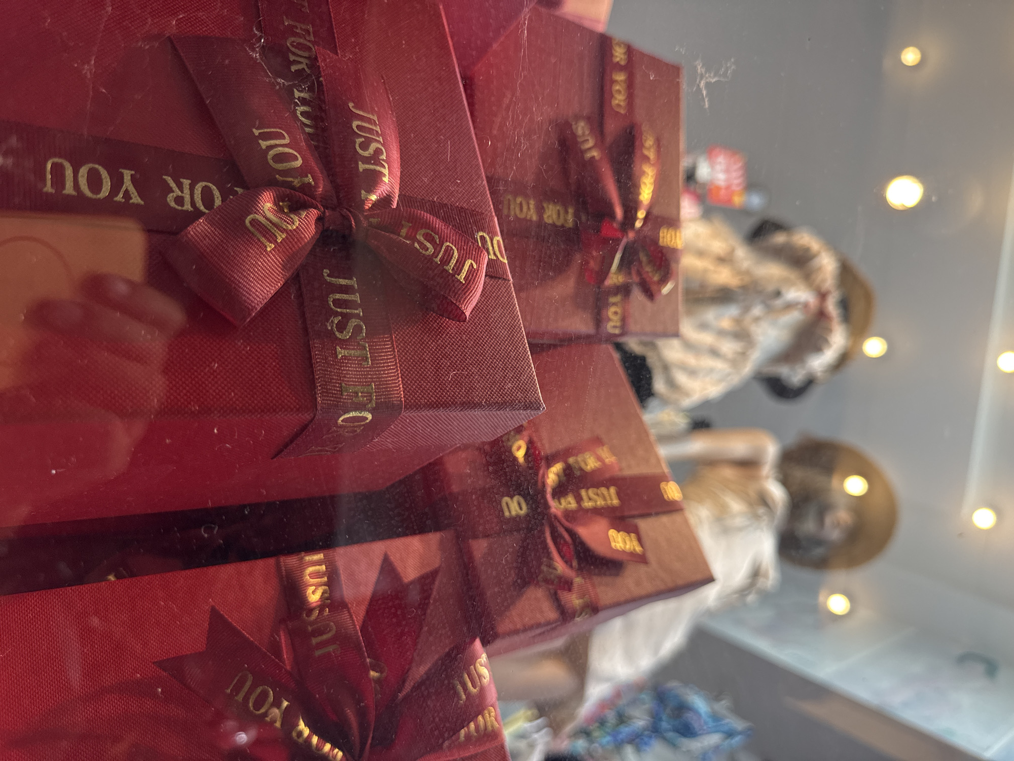 In Nha Trang Ward, Vietnam, a close-up shot focuses on several red gift boxes with gold ribbons tied in bows. The ribbons are inscribed with the words "JUST FOR YOU" in gold lettering. The boxes are arranged in a way that suggests they are displayed in a shop window, as indicated by the reflections on the glass surface. In the blurred background, mannequins can be seen wearing hats and dresses, further reinforcing the retail setting. The lighting is bright, with several light sources creating bokeh effects. The overall impression is one of a gift shop or boutique.