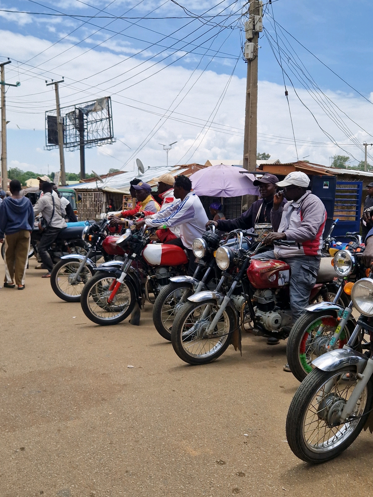 The image depicts a street scene in Kabong, Nigeria, with several men and motorcycles. A group of men are gathered around their motorcycles, which are parked in a row on a paved street. The men appear to be waiting or socializing. The environment is a typical urban street with buildings, utility poles, and overhead wires. The sky is partly cloudy. The overall atmosphere suggests a moment of pause or gathering in a busy area.