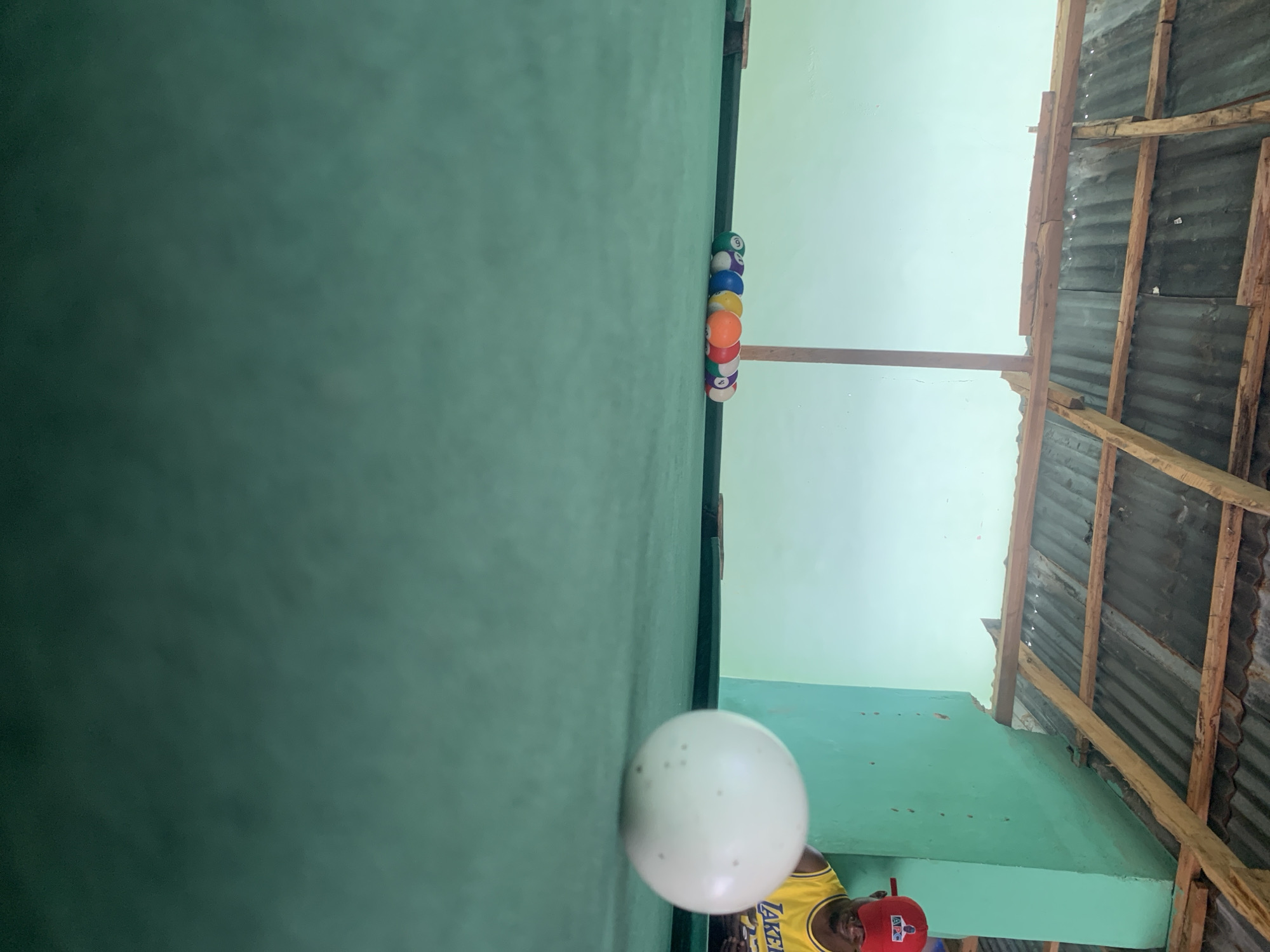 The image depicts a pool table covered in green felt, located in a structure with a roof made of corrugated metal and wooden beams. A rack of pool balls is positioned at the far end of the table. A white cue ball is in the foreground. A man wearing a yellow Lakers jersey and a red cap is partially visible in the lower center of the frame. The location is near New Stadium, Nigeria.