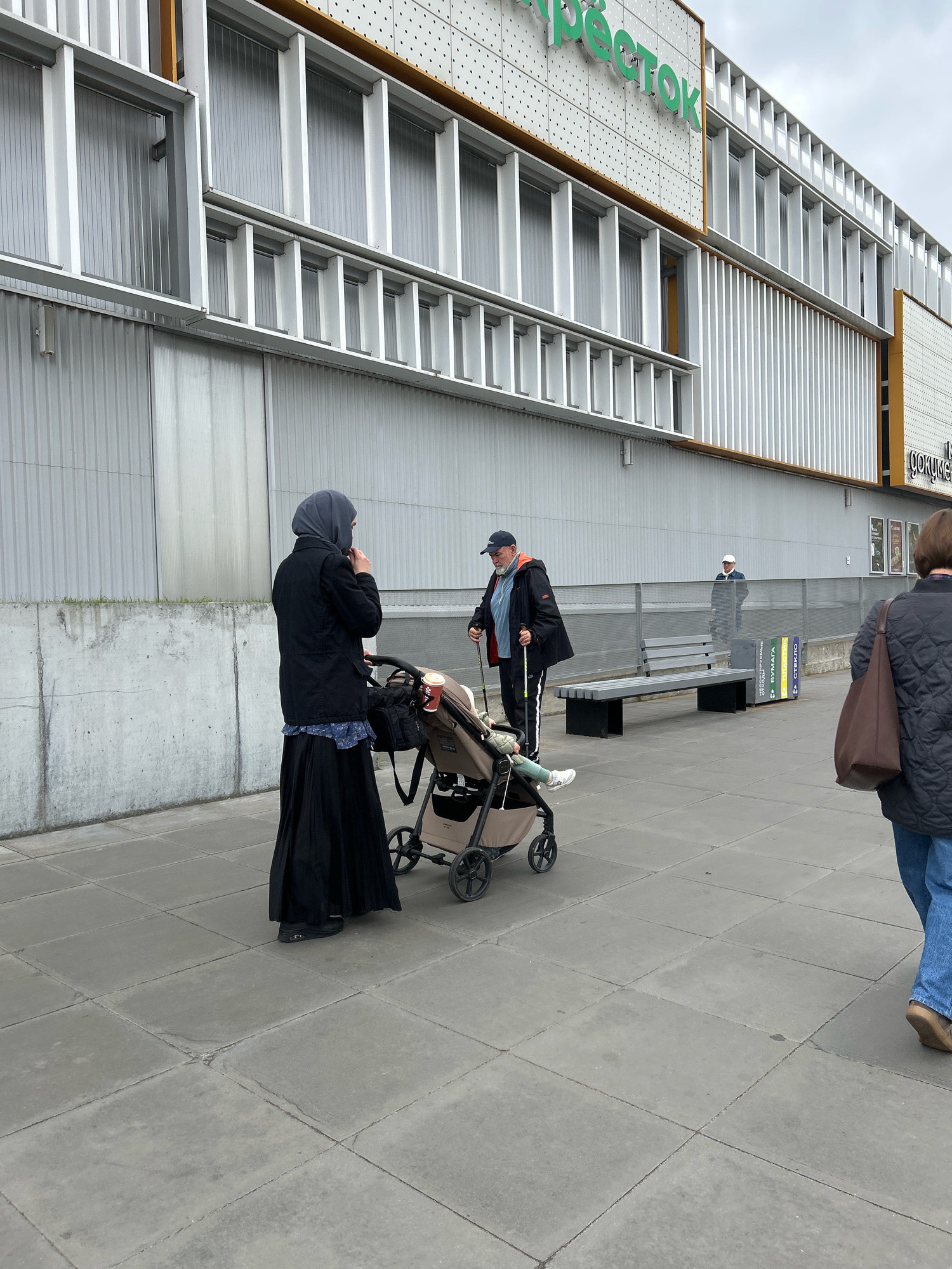 In Moscow, Russia, outside a shopping center with a sign that reads "Перекрёсток" (Perekrestok), two individuals are walking. A woman in a dark hijab and long black skirt pushes a baby stroller. Beside her, a man with a blue cap, black jacket with orange trim, and athletic pants uses walking poles. Further in the background, another man in a dark jacket and white cap stands near a bench and recycling bins. The ground is paved with concrete tiles.