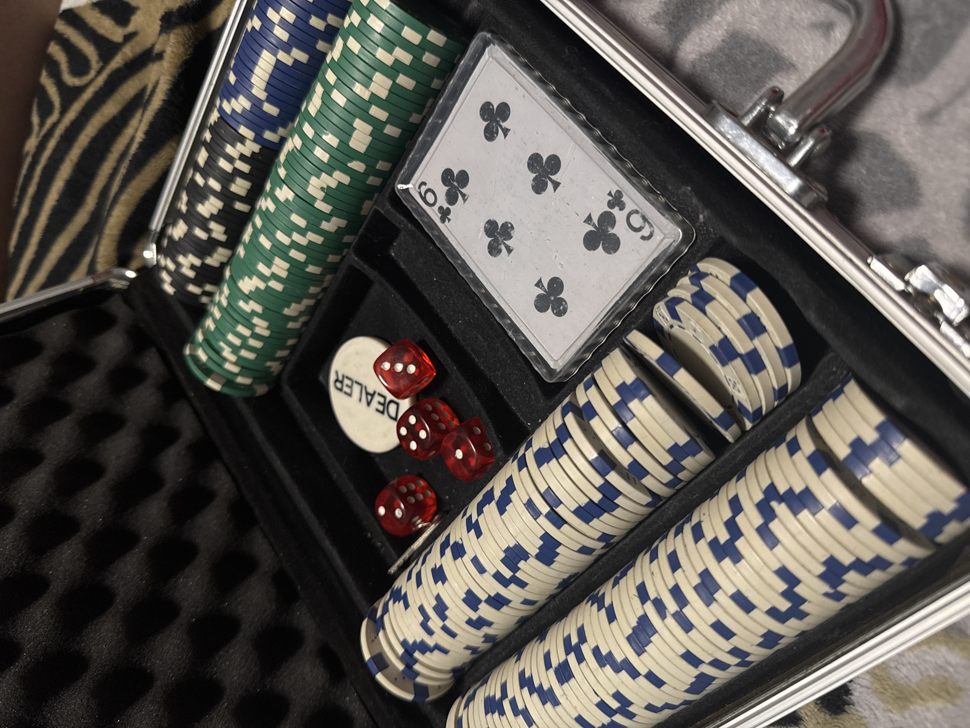 An open poker chip case contains stacks of blue, green, and black poker chips, a dealer button, red dice, and a six of clubs playing card. The case has a silver handle and is lined with black foam. The case is resting on a textured surface. There is no indication of this image's location being in the city of Izhevsk, Russia.