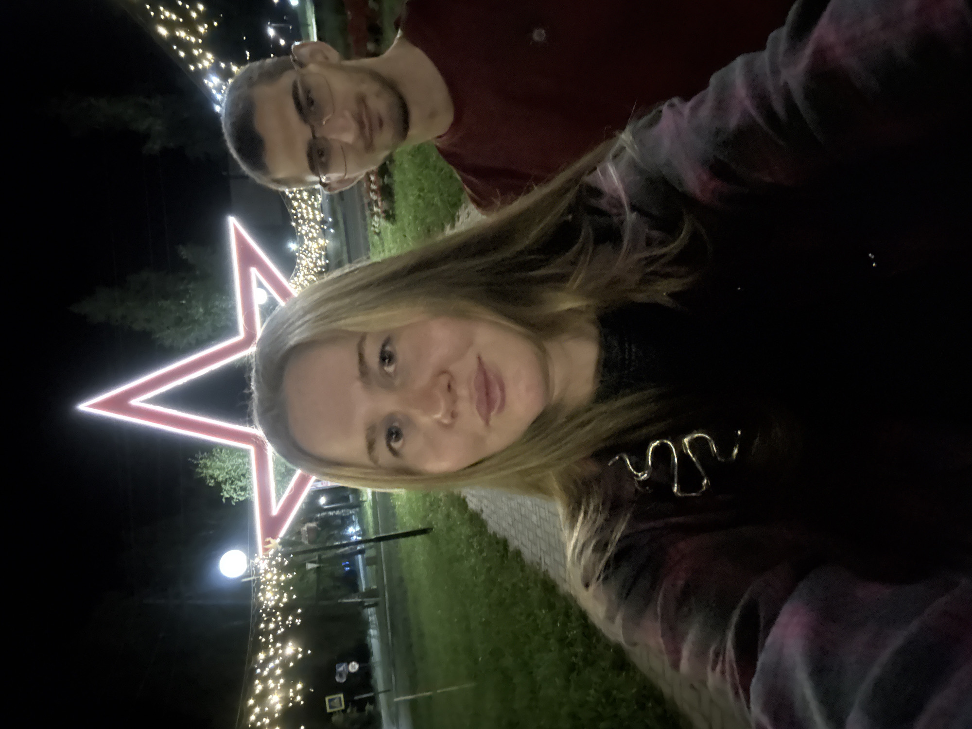 Two individuals, a woman and a man, are captured in a selfie taken at night. The woman, with blonde hair, is in the foreground and is wearing a dark, tie-dyed garment. A silver, snake-shaped hair accessory is visible in her hair. The man, with short dark hair and glasses, is positioned behind her and to her right. He is wearing a red t-shirt.
The backdrop features a large, illuminated red star outline, characteristic of decorative holiday lighting. Additional strings of warm white lights are draped in the background, creating a festive atmosphere. The scene appears to be outdoors, possibly in a park or public space, with a paved walkway and grassy areas visible. Trees are present in the background. The presence of streetlights and signage suggests an urban or suburban setting. The image is believed to have been taken in the city of Mozhga, Russia, indicated by contextual information.