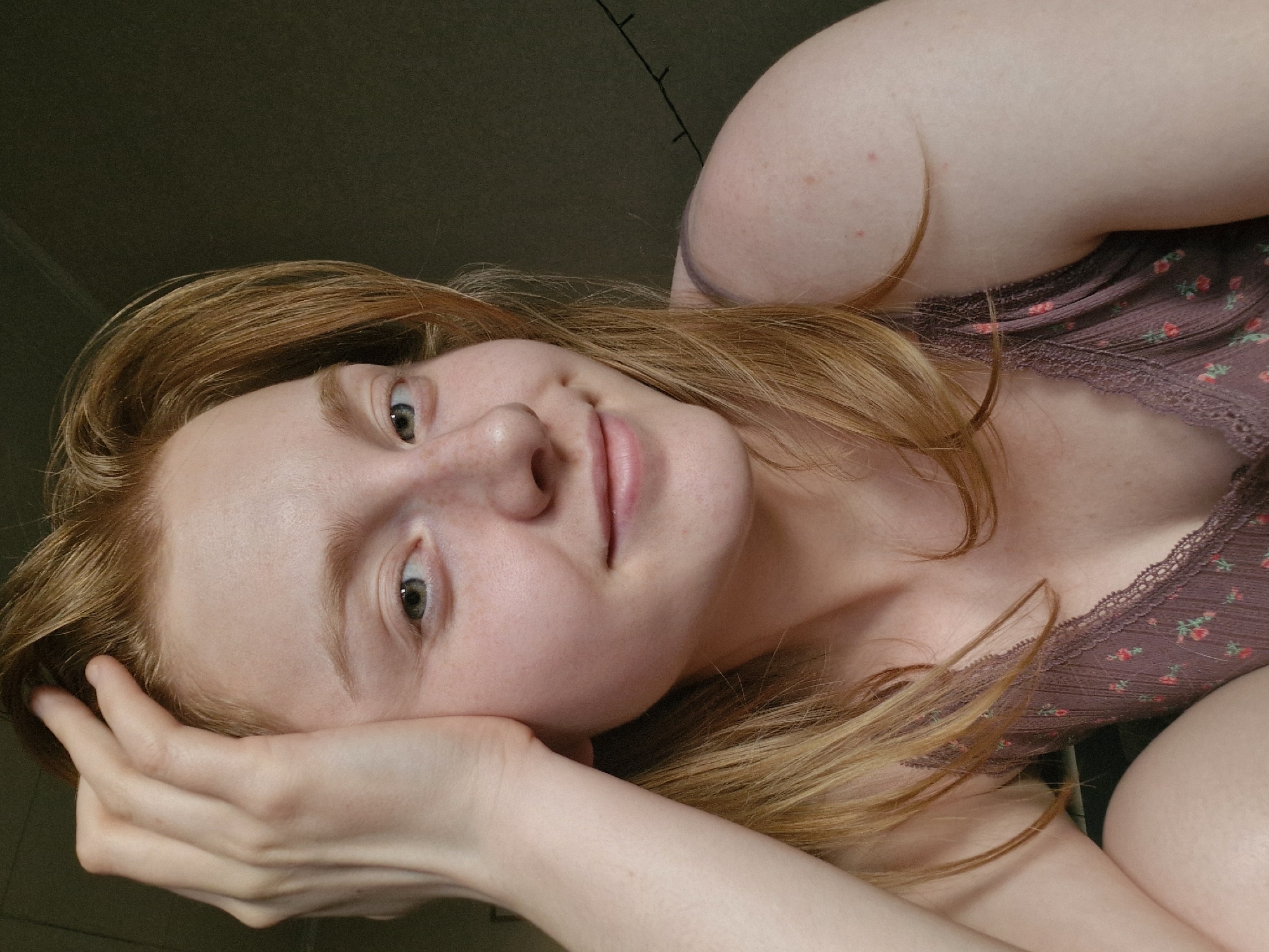 A close-up, portrait-style image shows a woman lying on her side. She has light skin, freckles, and reddish-blonde hair. Her right hand is supporting her head. She is wearing a brown top with a floral pattern and lace trim. The background is dark and out of focus. There is no indication of the image being taken in Yekaterinburg, Russia, or any city.