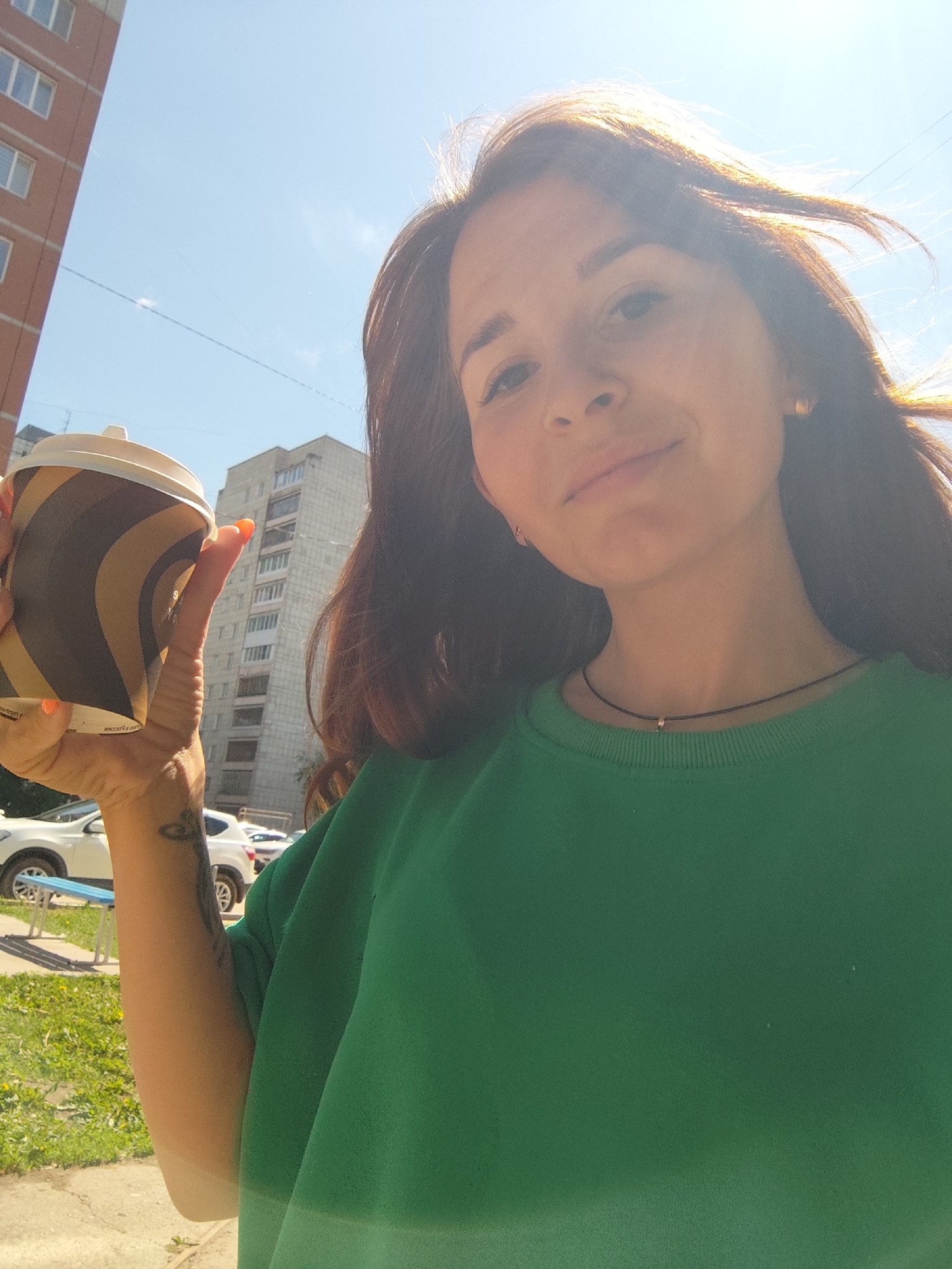 A young adult female is depicted in a brightly lit outdoor selfie, holding a disposable coffee cup with her right hand. She has long, dark hair, a fair complexion, and wears a plain green long-sleeved top. A thin, dark choker necklace with a small metallic charm is visible around her neck. Her fingernails are painted orange, and a dark tattoo is present on her inner right forearm. The coffee cup exhibits a spiral pattern in dark brown and light brown/tan colors and has a white lid. Partially visible text in Cyrillic is on the cup.
The background features an urban environment under a clear blue sky with faint wispy clouds. On the left, a tall, multi-story reddish-brown brick building with white-framed windows is visible. In the center-right background, another multi-story, grey concrete residential building with numerous windows is present. Several white cars are partially visible parked in the lower-left, adjacent to a patch of green grass with yellow dandelions. Thin overhead wires stretch across the sky. Strong, direct sunlight causes significant lens flare across the subject's face and hair. The architectural style of the buildings and the Cyrillic text on the cup are consistent with a location in the city of Chaikovsky, Russia.