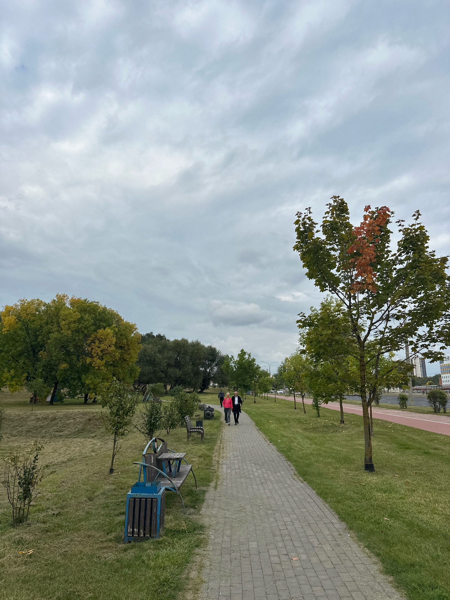 The image depicts a park path with a paved walkway in Minsk, Belarus. A couple is walking away from the camera on the path, with a man in a suit and a woman in a pink jacket. To the left of the path is a grassy area with trees, some of which are displaying autumn colors. There are also several park benches and a blue trash bin. To the right of the path is a red bike lane, and beyond that, a road with some buildings visible in the distance. The sky is overcast with gray clouds.