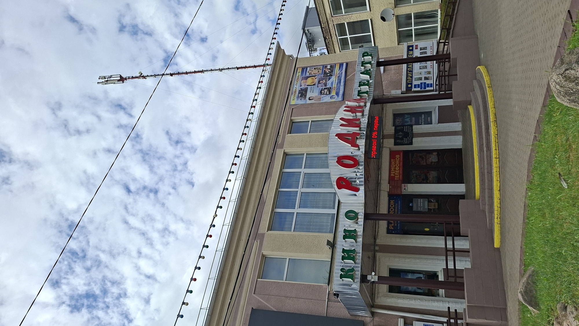The image shows the exterior of the "Rodina" cinema in Pastavy, Belarus. The cinema is a two-story building with a light-colored facade. Above the entrance, a sign reads "КИНО РОДИНА ТЕАТР" (Cinema Rodina Theater). There are posters displayed on the building, including one for a television channel and another advertising movie showtimes. Red and green lights are strung across the top of the building. A tall cell tower is visible in the background. The entrance to the cinema features a set of stairs with yellow markings. The area in front of the cinema is paved, and there is a grassy area with rocks in the foreground. The sky is cloudy.