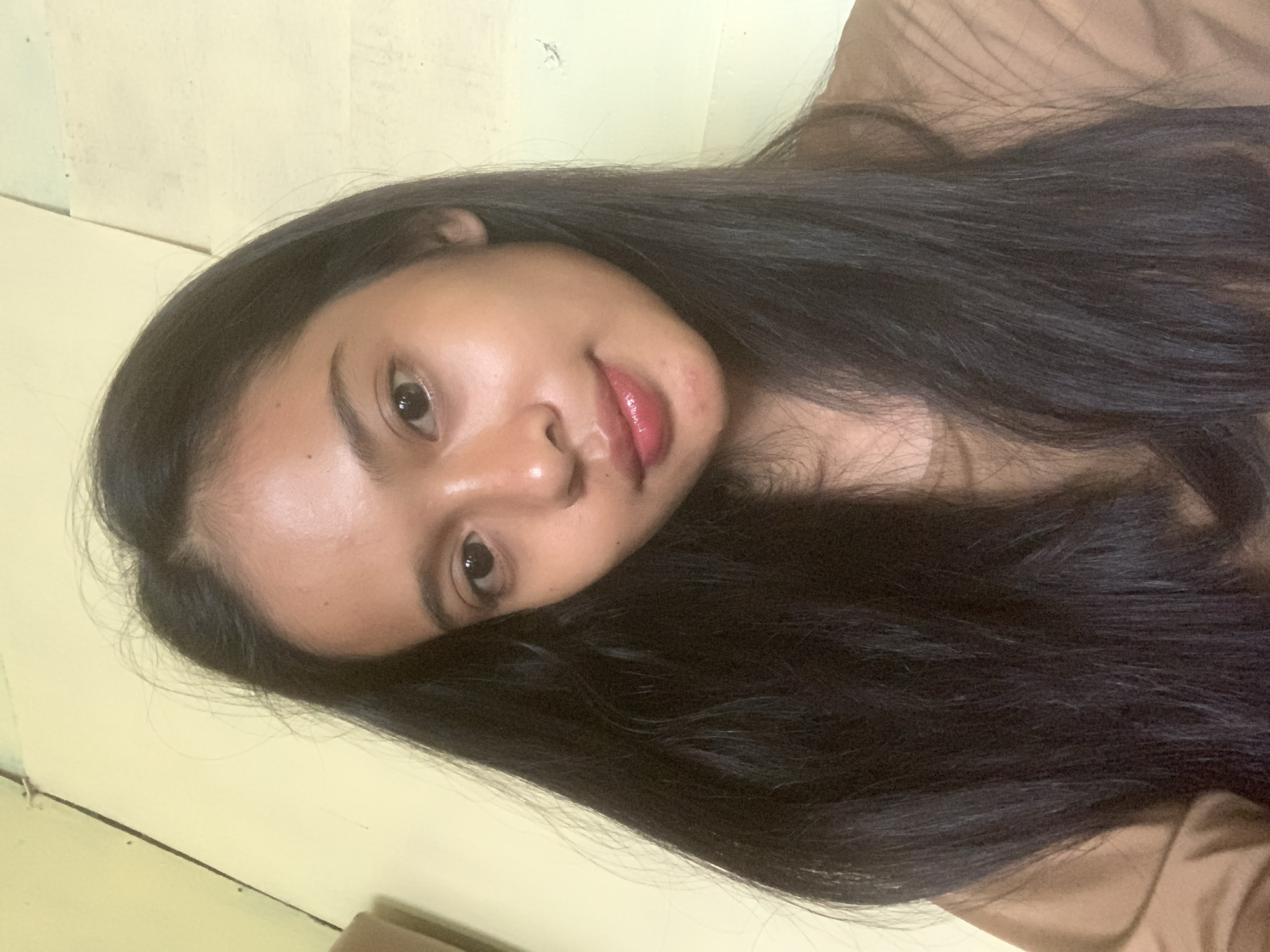 A young individual, identified as female, with long, straight, dark hair parted centrally, is depicted in a headshot taken indoors in Sungai Meriam, Indonesia. The subject's face is oriented directly towards the camera, displaying a neutral expression with a slight smile. The skin tone is medium, with visible highlights indicating natural sheen on the forehead, nose, and cheeks. The eyes are dark, looking straight into the lens. The lips are full, coated with a glossy, reddish-pink product, and minor skin irregularities are present on the chin. The subject is wearing a light brown, possibly tan, crew-neck top, visible across the shoulders and upper chest. The background consists of a light yellow-cream colored wall exhibiting irregular texture, subtle discoloration, and faint vertical lines on the right side. A darker, indeterminate object is partially visible along the left edge of the frame. The lighting is uniform, illuminating the subject's face without strong shadows.
