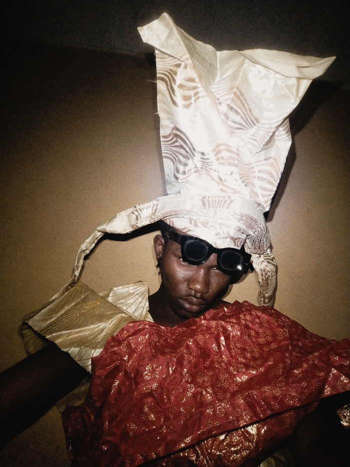 A person wearing sunglasses and a towering, patterned headwrap poses in front of a wall. They also wear a gold-colored garment on their shoulder and a reddish-gold patterned cloth is draped in front. The person has dark skin and appears to be gazing downward. The lighting is dim, creating a somewhat theatrical or dramatic mood.