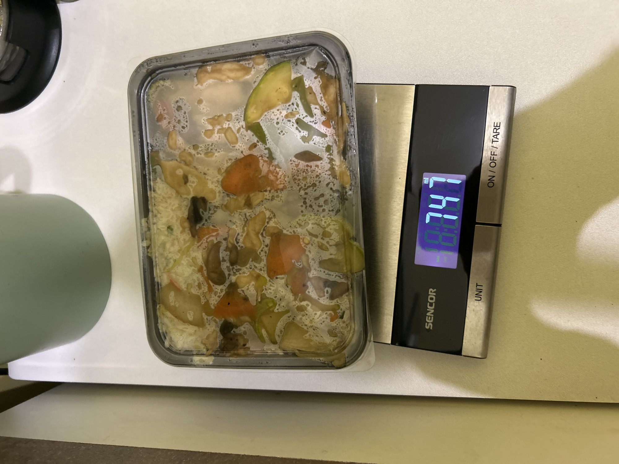 A plastic container with food sits on a digital scale. The scale reads 747. The container is clear and rectangular, with a gray rim. The food inside appears to be a mixture of rice, vegetables, and broth or sauce. The vegetables appear to be carrots, zucchini, and mushrooms. The scale is a Sencor brand. The background is a light cream color.
It is impossible to determine from this image whether the location is Sofia, Bulgaria.