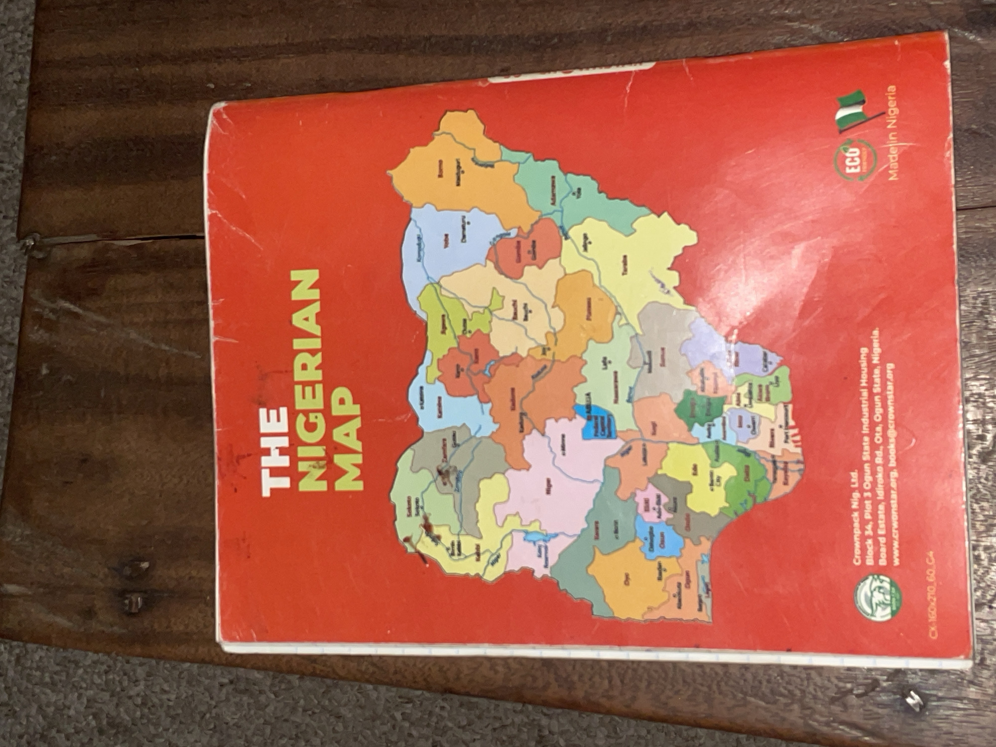 The image shows the back cover of a map of Nigeria. The title "THE NIGERIAN MAP" is prominently displayed in large, lime green letters on a red background. Below the title is a colorful political map of Nigeria, divided into states with their names labeled. There are also text details at the bottom of the cover. These include the publisher's information: "Crownpack Nig. Ltd.", "Block 34, Plot 3 Ogun State Industrial Housing Board Estate, Idiroke Rd., Ota, Ogun State, Nigeria.", and website/email addresses: "www.crwonstar.org, books@crownstar.org". There is also a circular "ECO" logo with a leaf and an image of Nigeria's flag, followed by the text "Made in Nigeria". The map itself does not specifically highlight the city of Jalingo; it shows Jalingo as a point within the Taraba State of Nigeria. The overall scene is a flat lay of the map cover on a textured wooden surface.