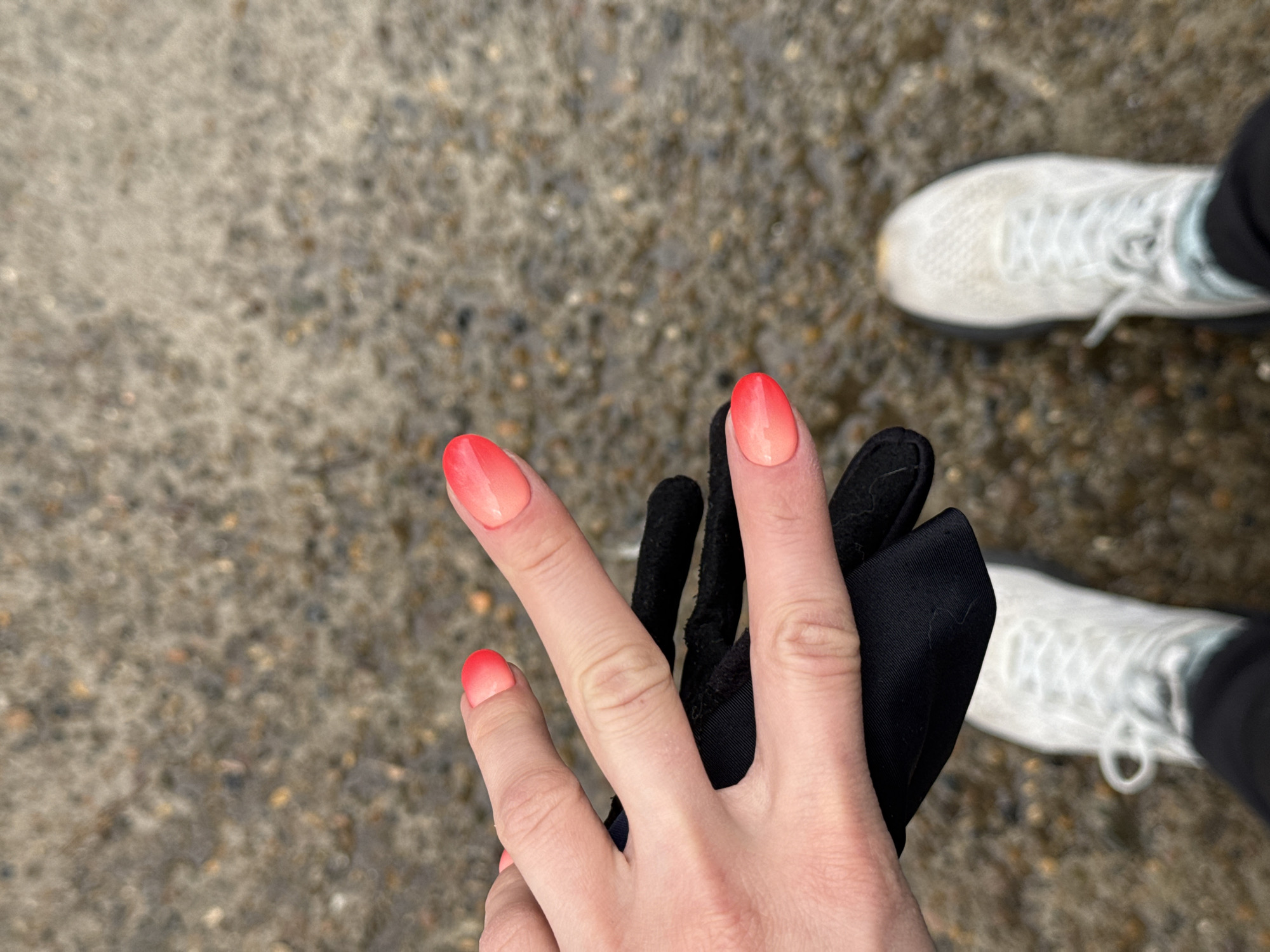 An overhead shot captures a person's left hand against a backdrop of damp, speckled gray pavement. The hand features short, oval-shaped nails, each painted with a striking red-to-light-pink ombre design. The person's fingers are slightly spread, and they are holding or partially wearing a black glove made of a smooth, possibly synthetic, fabric. In the background, the front of a white athletic shoe with white laces is visible on the right side, along with the toe of another white shoe in the upper right. The pavement appears wet or damp, indicating recent rain or humid conditions, and the diffused light suggests an overcast daytime. The scene is an outdoor setting, likely a path or sidewalk in Chaikovsky, Russia, implying the person is walking or standing in potentially cool or wet weather. No text is visible.