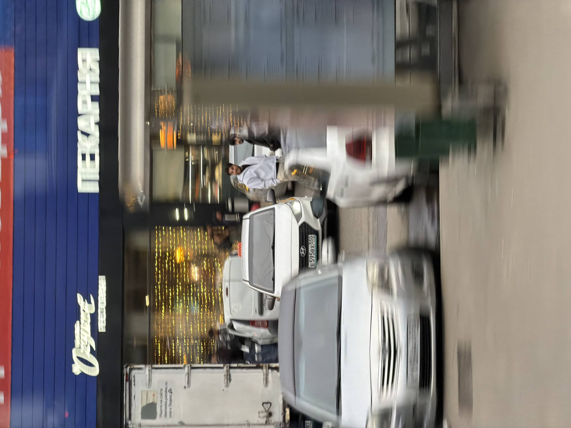 Here is a description of the image:
The image appears to be taken on a city street in an unknown location. There's a building visible on the left with signage in a script style, possibly storefront advertising, and a large sign that reads "ПЕКАРНЯ" vertically on the building's blue facade. String lights are visible in the building's window, suggesting an interior space like a shop. The street scene is busy with parked cars. The license plate of a vehicle is partially visible, showing "E595BX150". There are pedestrians around the vehicles. The overall impression is that of an urban environment with commercial activity.