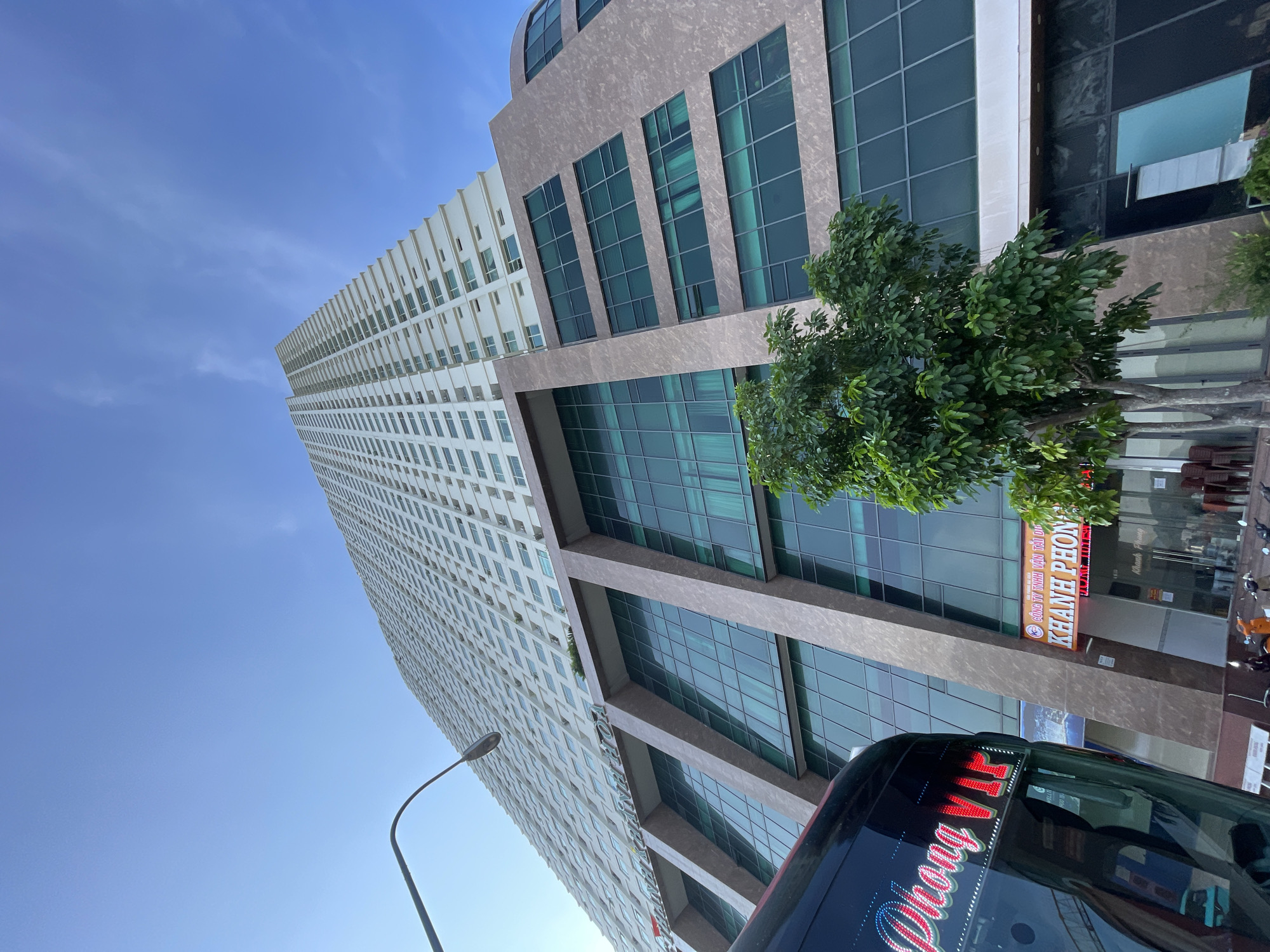 A tall, white, multi-story building with numerous windows dominates the frame, reaching towards a clear blue sky. The building's lower levels feature a facade with large, diagonal brown and beige panels and green-tinted windows. A portion of a black bus with red and white lettering that reads "Phuong VIP" is visible in the lower left corner. In front of the building's ground floor entrance, there is a small tree with dense green foliage and a sign that reads "CÔNG TY TNHH VẬN TẢI DU LỊCH KHANH PHONG". Several chairs are stacked outside the entrance, and some motorcycles are parked on the sidewalk. The scene appears to be a commercial or residential building in an urban environment.