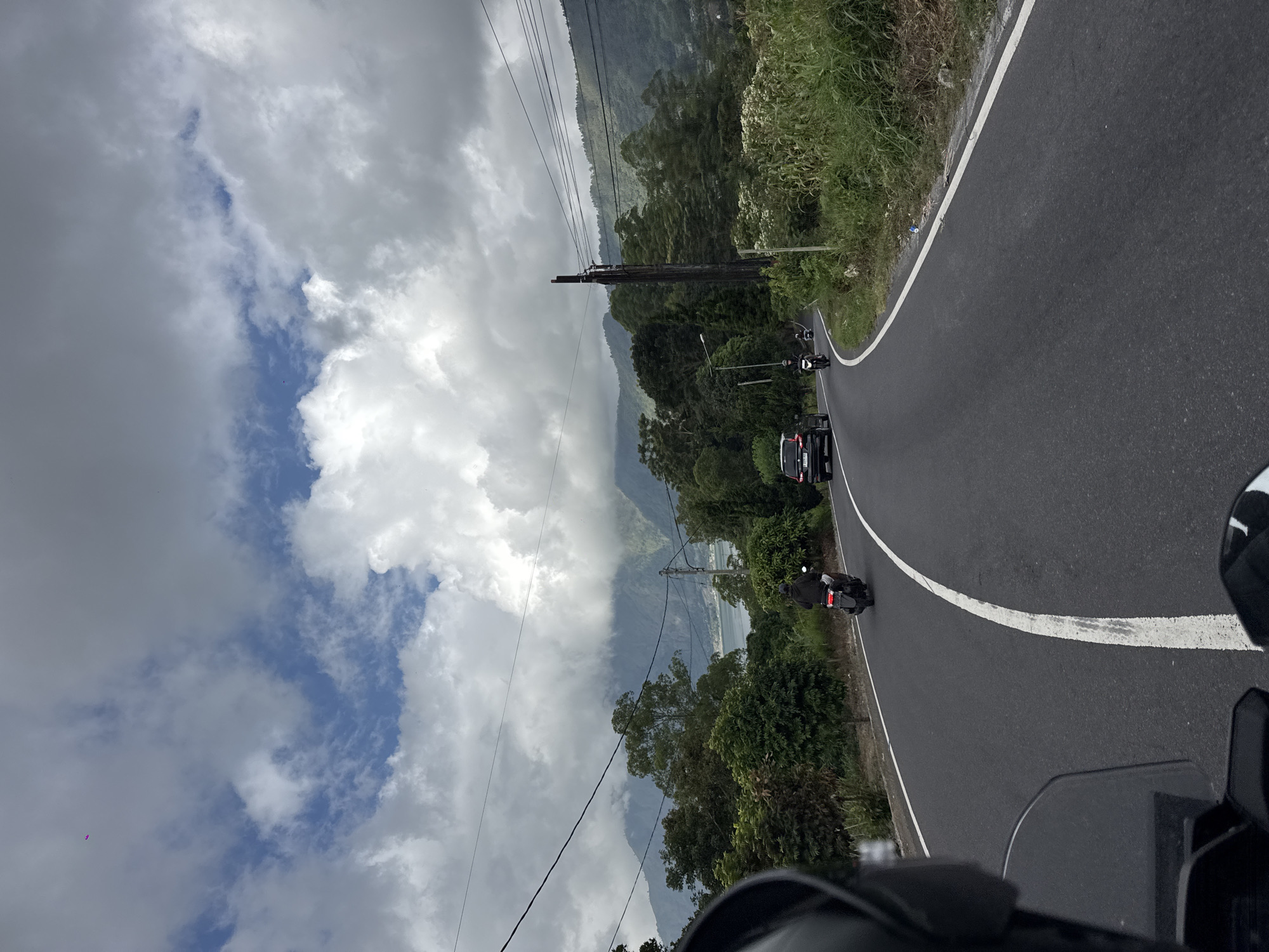 The image depicts a scenic road in Kintamani, Indonesia, with lush green mountains and a cloudy sky. The road is paved and curves to the left, with a white line marking the lane. There are several vehicles on the road, including a black car, two motorcycles, and a motorcycle rider in the foreground. The mountains in the background are covered in mist, and there is a body of water visible in the distance. The vegetation on the sides of the road is dense and green, with a variety of trees and plants. The overall scene suggests a beautiful and somewhat dramatic landscape, possibly during or after a rain shower.