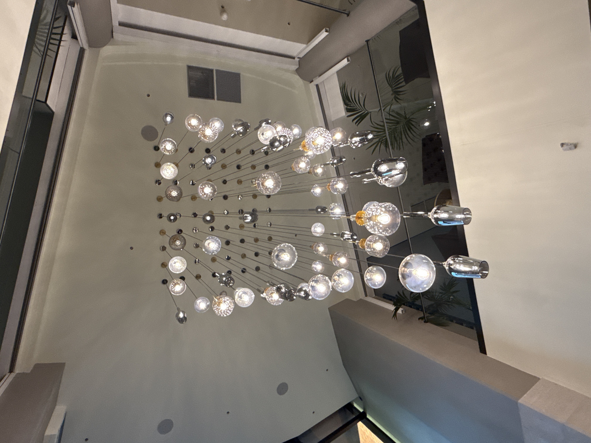 The image shows a modern chandelier with multiple glass spheres suspended from thin metal rods, hanging in a large open space. The chandelier appears to be a central feature of the architecture. The space has high ceilings and neutral-colored walls, and large glass windows with views of what appear to be indoor plants. There is no evidence to support the claim that it is located in Moscow, Russia.