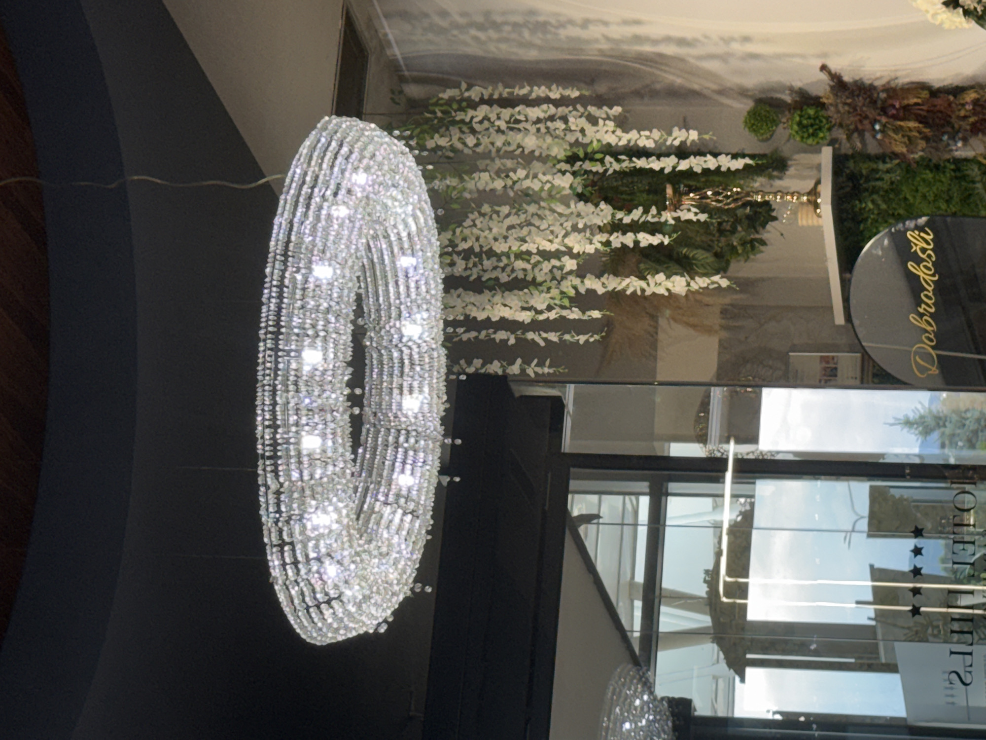 The image shows an ornate crystal chandelier hanging from a dark ceiling. Below the chandelier, to the right, is a decorative display featuring cascading white artificial flowers, a golden candelabra, and lush greenery. In the foreground, a circular black sign with golden cursive text reads "Dobrodošli". To the left, a glass door with "HOTEL HILLS" and three stars is visible, suggesting the location is Hotel Hills in Ilidža, Bosnia and Herzegovina. The environment appears to be an indoor space, possibly a hotel lobby or reception area.
