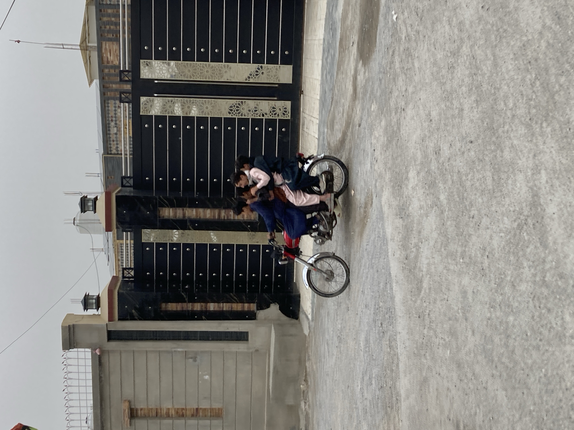 The image depicts a motorcycle carrying multiple people on a paved street in what appears to be a residential area within Quetta City Tehsil, Pakistan. A large, ornate gate stands behind the motorcycle, suggesting the entrance to a property. The building next to the gate is of light concrete construction. The day appears overcast. There are three discernible people riding on the motorcycle. The rider and passengers are unidentified.