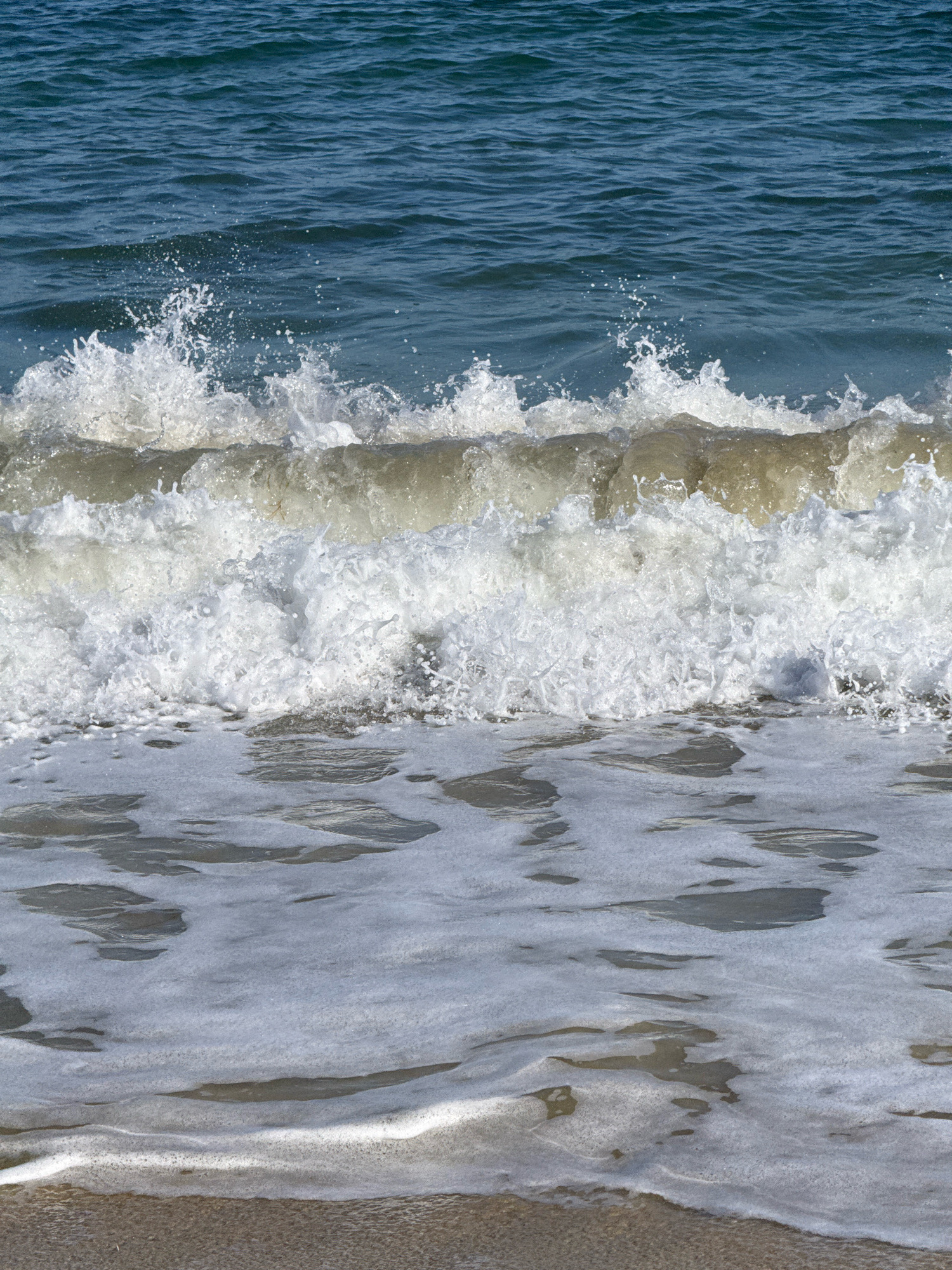 The image shows a close-up, eye-level view of a breaking wave on a sandy beach. The water is a deep blue, with a white foamy crest on the wave. The wave is in mid-break, with water splashing and creating a spray of white. The sand is a light brown color, and the water is reflecting the sunlight. The scene is set in the city of Kiten, Bulgaria, likely on one of its beaches. There are no people or other objects visible in the image.