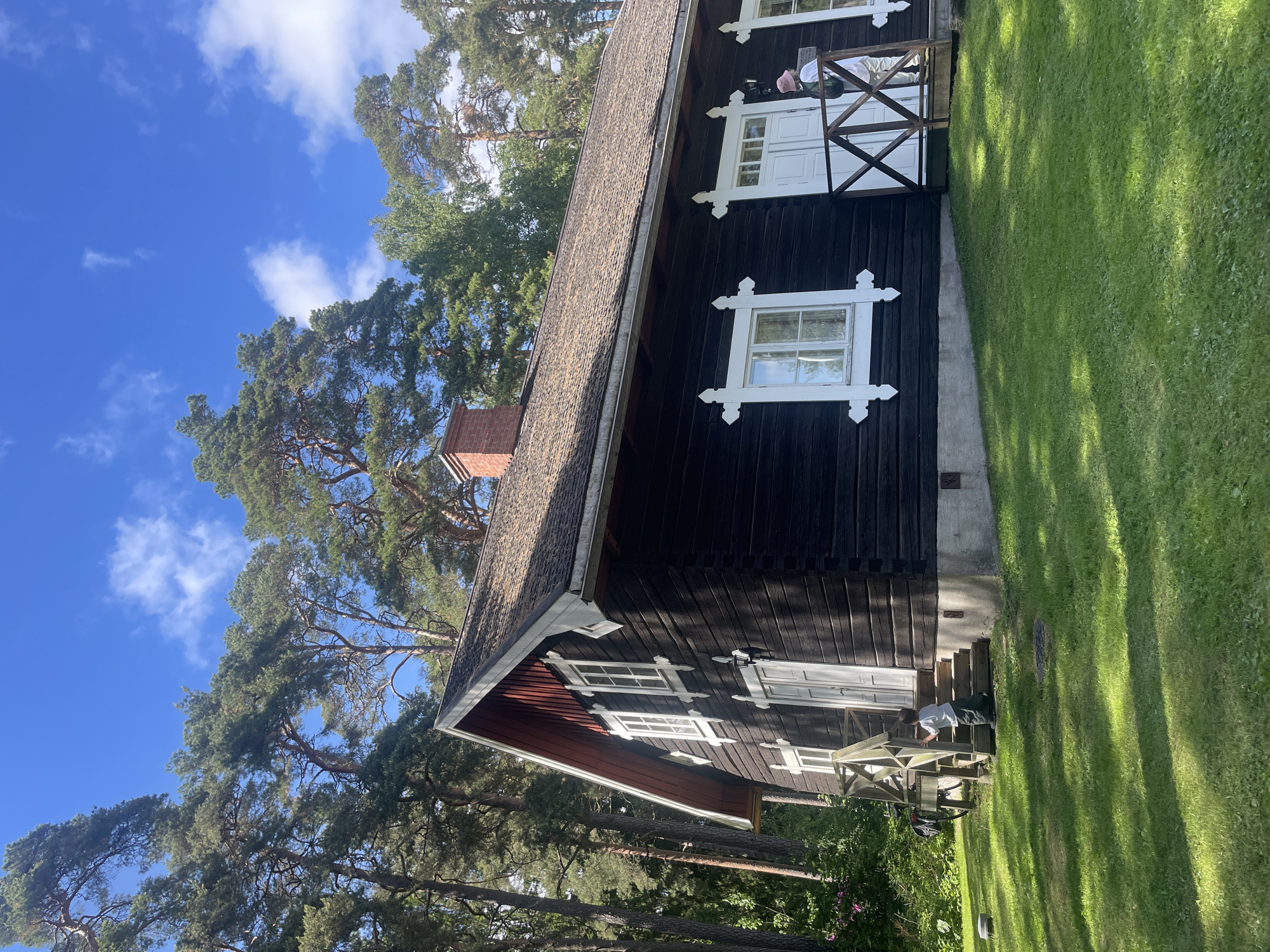 The scene depicts a traditional dark wooden house with white window and door trim, situated in a grassy, forested area under a clear blue sky in Karlstad, Sweden.
The house features a gabled roof with dark brown shingles and red-painted eaves on the visible gable end. A red brick chimney is visible on the roof towards the right side. The exterior walls are constructed from dark-stained logs, featuring prominent white decorative cross-shaped elements below and above the windows and doors. Multiple white-framed windows, some with multiple panes, are present. Two white double doors, each accessed by small wooden porches with railings, are visible. The house rests on a light-colored foundation, possibly concrete.
Two individuals are present near the house. On the left side, a small child wearing a light-colored top and dark trousers stands on the wooden steps of a porch, holding the railing. To the right, an adult wearing light-colored clothing, a pink bucket hat, and a dark backpack stands on a separate wooden porch, facing the house entrance. A small, dark rectangular sign with white text is affixed to the house wall near this adult.
A bicycle with a rear rack and bag is partially visible near the steps of the left porch. On the grass to the far left, a low, circular, light-colored object is present. A circular manhole cover is embedded in the grass in front of the right side of the house.
The surrounding environment consists of numerous tall pine trees, their green foliage extending high into the blue sky, which contains scattered white cumulus clouds. The foreground is covered by a vibrant green lawn exhibiting distinct patterns of sunlight and shadow, indicating a sunny day. The ground appears to slope gently upwards towards the house.