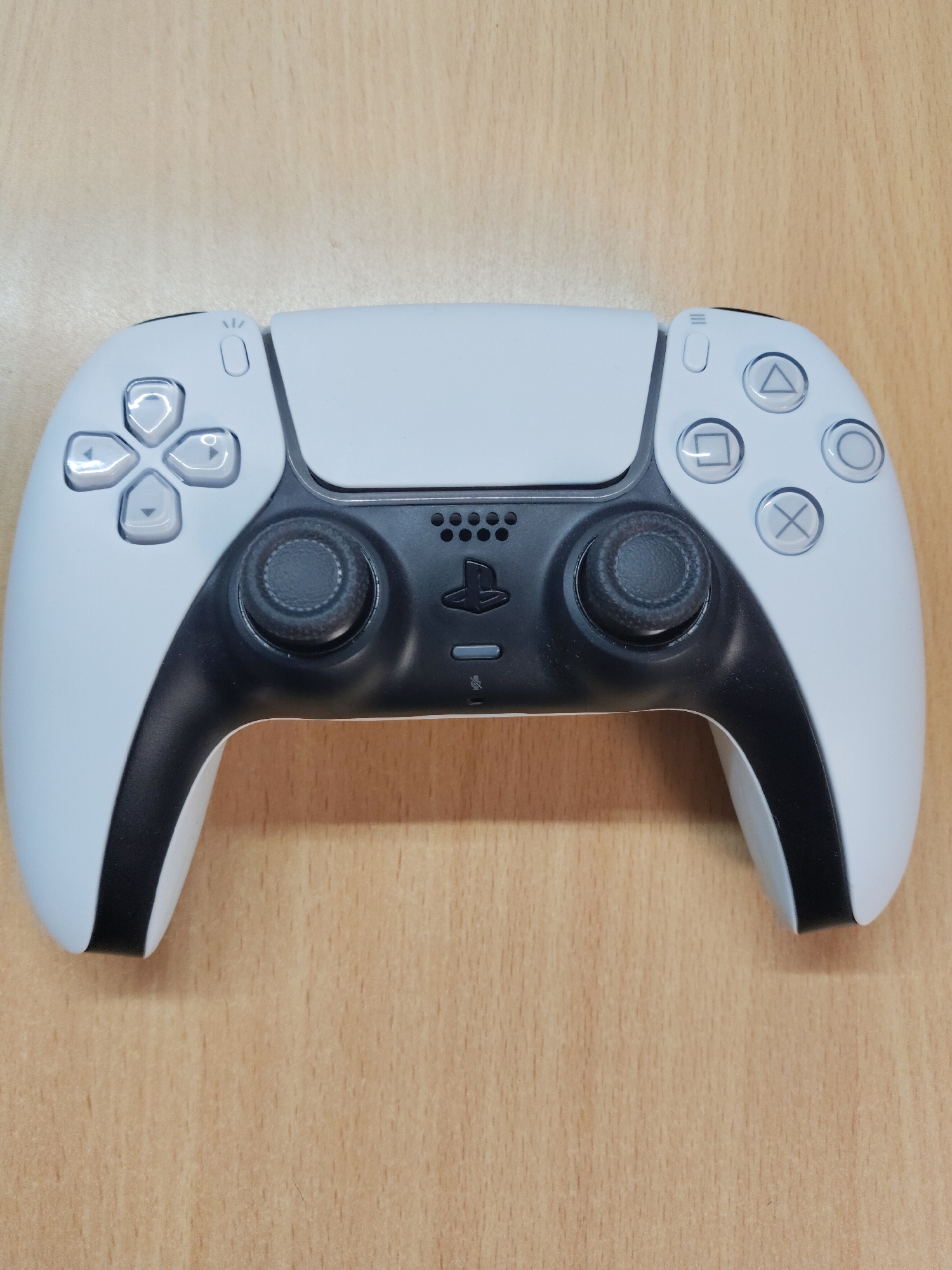 A PlayStation 5 DualSense controller sits on a light brown, possibly wood-grain, surface. The controller's white and black color scheme is visible, with its directional pad, analog sticks, face buttons, and PlayStation logo clearly displayed. The controller is positioned in the center of the frame. The setting is an indoor environment. There is no indication that the image was taken in Perm, Russia.