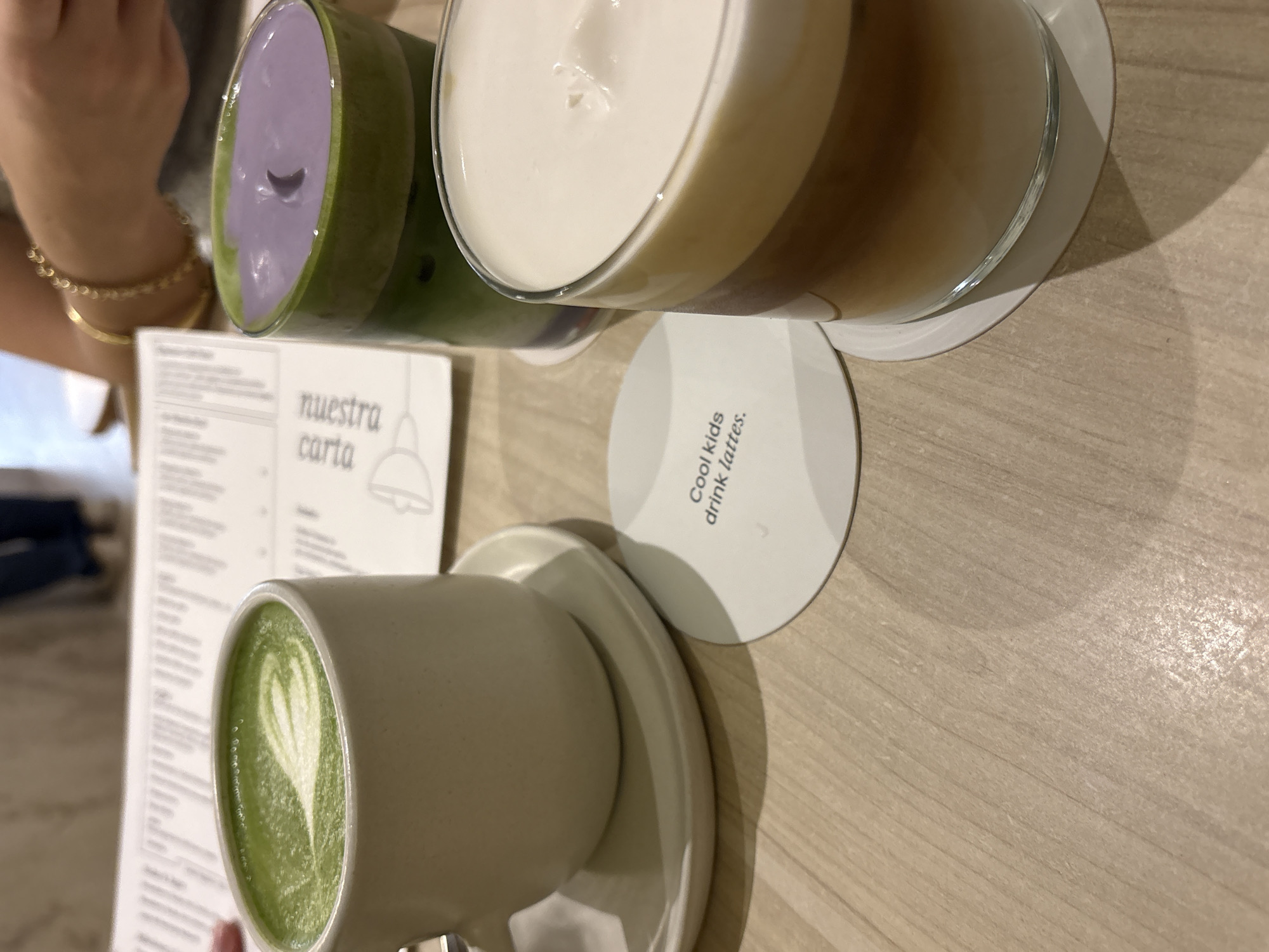 The image depicts a close-up of three beverages on a light-colored wooden table. In the foreground to the right, a glass contains a layered drink with shades of brown and white foam. To the left, a white mug holds a green matcha latte with intricate foam art. Behind the mug, a clear glass contains a layered drink with a green base and a purple topping. A white circular coaster with the text "Cool kids drink lattes." is placed on the table between the mug and the glass with foam. In the background, a menu is partially visible, and a person's arms, wearing a gold bracelet, are resting on the table. The setting appears to be an indoor cafe. The image does not provide any specific information about the city of San Isidro, Peru.