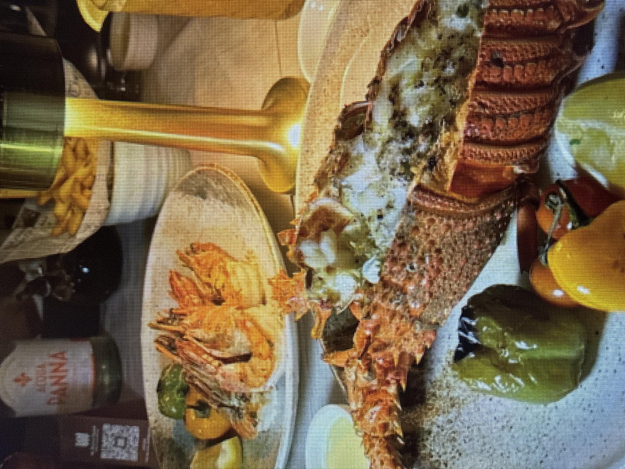 The image shows a close-up view of a table setting with a meal. In the foreground, a large, cooked lobster is presented on a white plate, split lengthwise and seasoned with herbs. To the left of the lobster, a green bell pepper and other vegetables are visible, along with a small bowl containing a yellow sauce. In the background, a platter of grilled shrimp is also on display, along with a bottle of San Pellegrino water and a basket of fries. A golden lamp with a cylindrical shade illuminates the scene. The setting appears to be a restaurant, though the specific location is unknown.