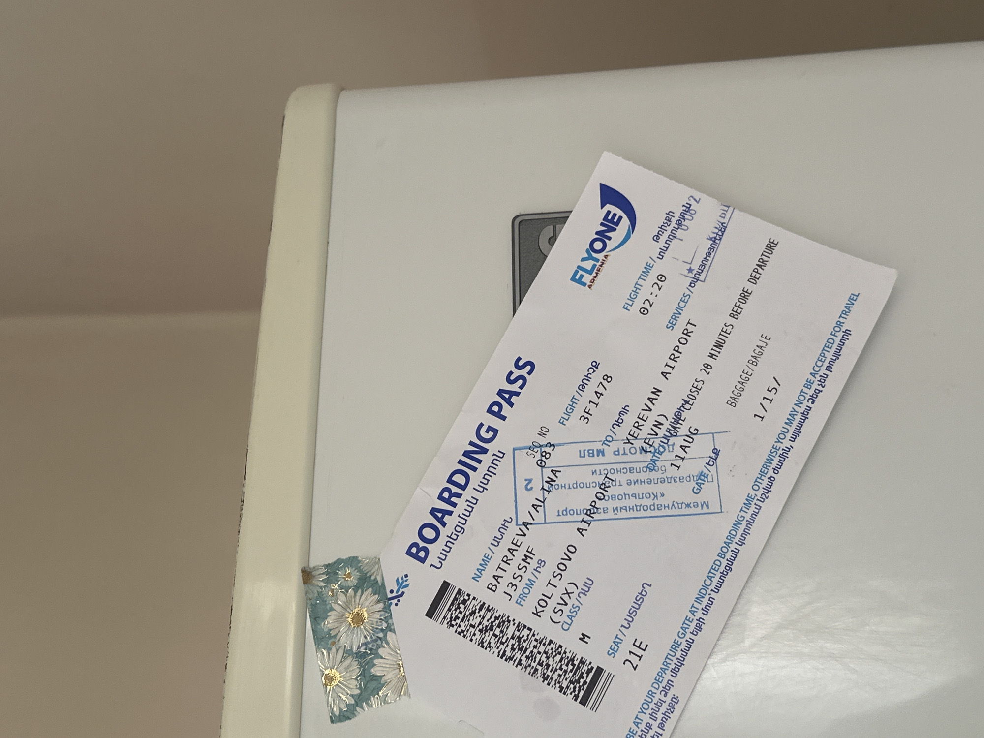 The image shows a close-up of a FlyOne Armenia boarding pass, possibly on a white refrigerator. The boarding pass has a barcode on the left side and text in English and Armenian. It lists passenger information, flight details, and departure information. Notably, it shows the destination as "YEREVAN AIRPORT (EVN)" and the origin as "KOLTSOVO AIRPORT (SVX)". The date is "11 AUG", and the flight time is "02:20". The text at the bottom indicates "BE AT YOUR DEPARTURE GATE AT INDICATED BOARDING TIME. OTHERWISE YOU MAY NOT BE ACCEPTED FOR TRAVEL". The airline logo "FLYONE ARMENIA" is visible on the right side. There is also a small piece of decorative tape with a floral pattern on the upper left corner of the boarding pass. The background is a plain, light-colored surface, suggesting the boarding pass is placed on an appliance. No people or specific environmental details related to Gyumri, Armenia are depicted in the image.
