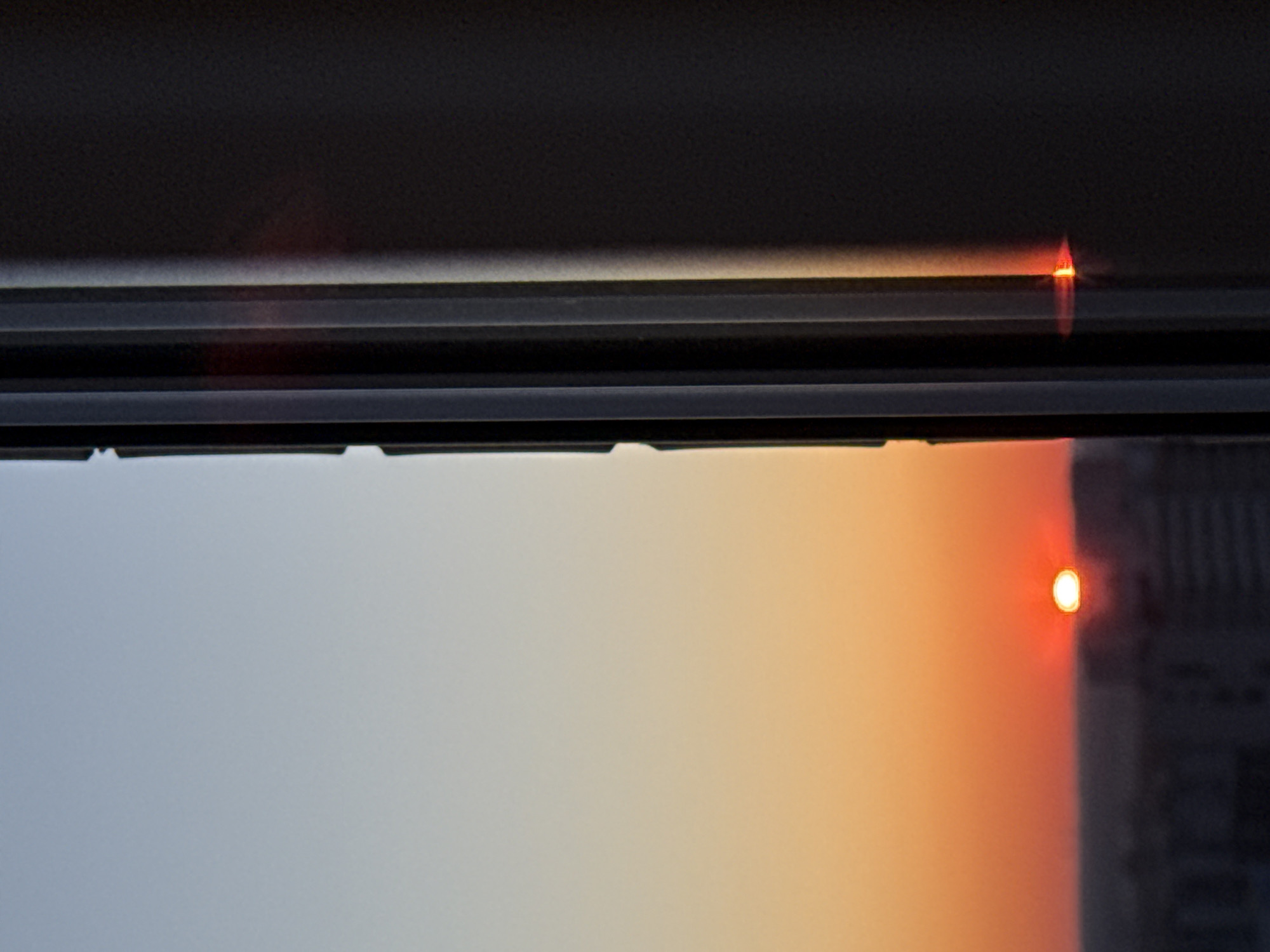 The image shows a partial view through a window, framed by the window's dark-colored structure. Outside, the sky transitions from a pale blue-gray to an orange hue, indicative of either a sunrise or sunset. A blurred, dark structure is visible on the right, with a bright, circular light illuminating its side. The scene appears to be an urban landscape, given the building visible through the window. The location is stated to be in Perm, Russia.