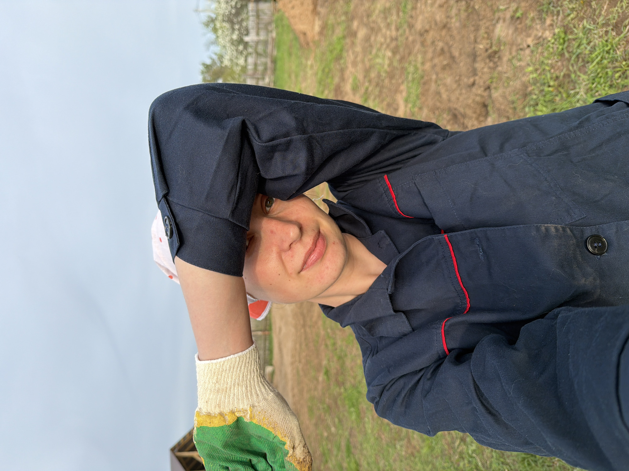 A person wearing a blue work shirt with red trim and a work glove on one hand is posing for a selfie outdoors. The person has light skin, short hair, and is wearing a white head covering. The background consists of a grassy area and some trees. It is not possible to confirm the location is Chaikovsky, Russia, based on the visual information provided.