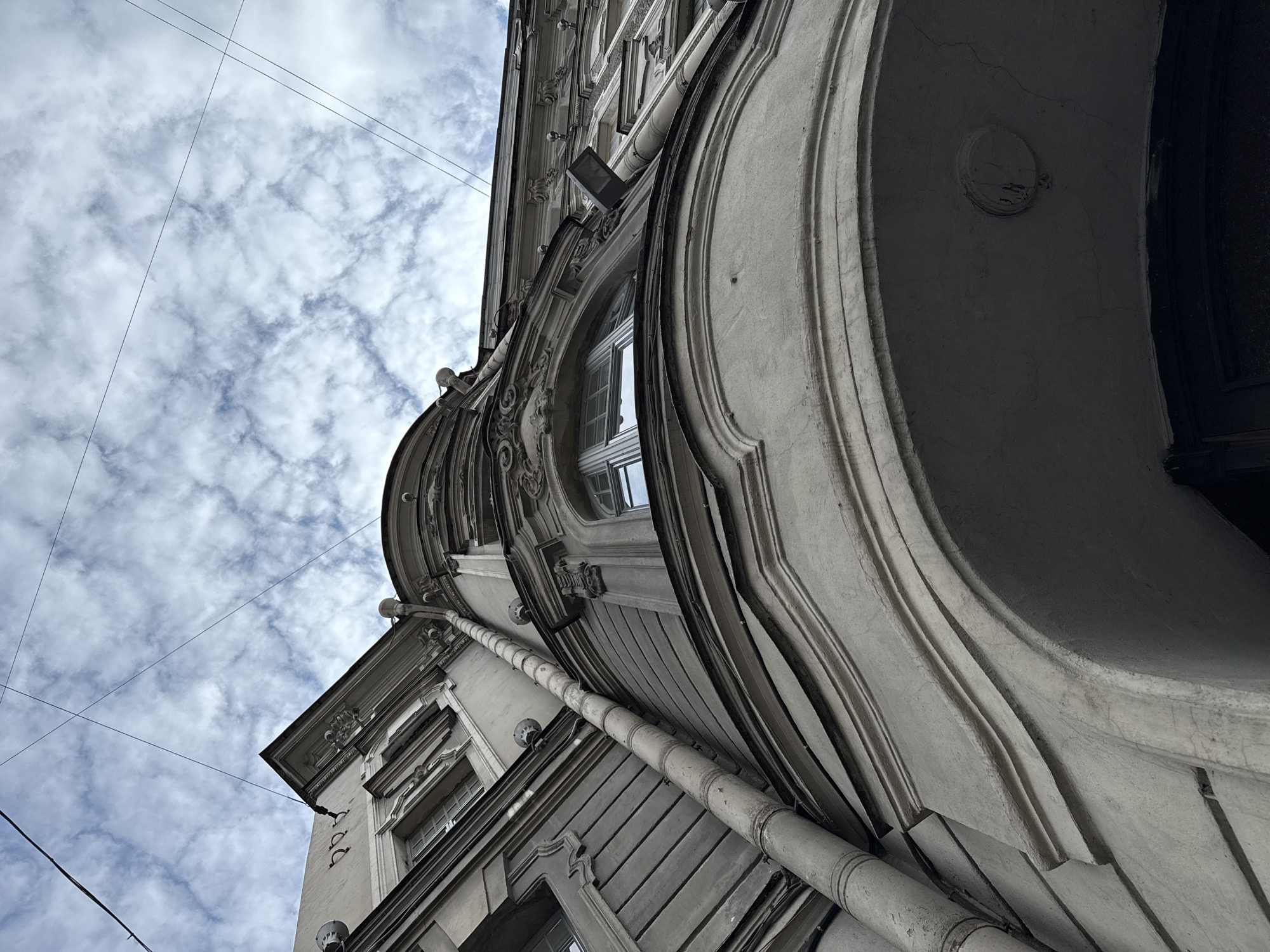 The image shows a low-angle view of an ornate building facade in Saint Petersburg, Russia. The architecture features classical elements such as decorative moldings, curved window frames, and a prominent drainpipe running vertically along the side of the building. The sky above is partly cloudy with wispy white clouds against a pale blue background. Several thin wires are visible crisscrossing the sky, likely utility or telephone lines. The building material appears to be stone or stucco, weathered and aged, with a greyish-white hue. The perspective emphasizes the grandeur and intricate detail of the building, with a focus on the upper levels and roofline against the sky. The scene is devoid of any people or discernible activity.