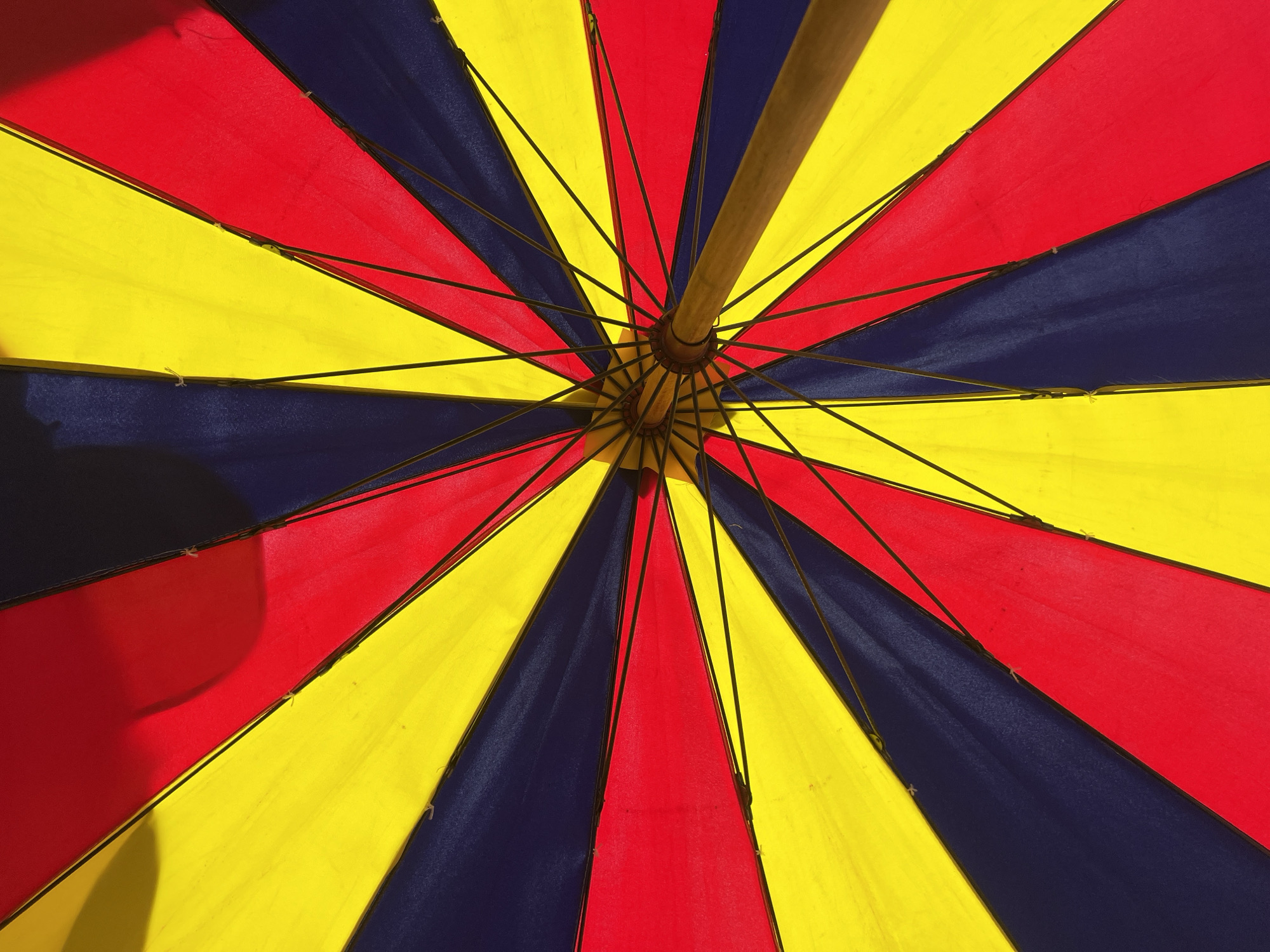 The image presents an upward view into the interior of a multicolored umbrella. The umbrella's canopy is segmented into alternating radial panels of yellow, red, and dark blue fabric. Black metal struts extend from a central wooden pole, supporting the fabric panels. The scene is brightly lit, highlighting the vibrant colors of the umbrella. The location in the city of Unknown, Unknown is not discernible from the image.
