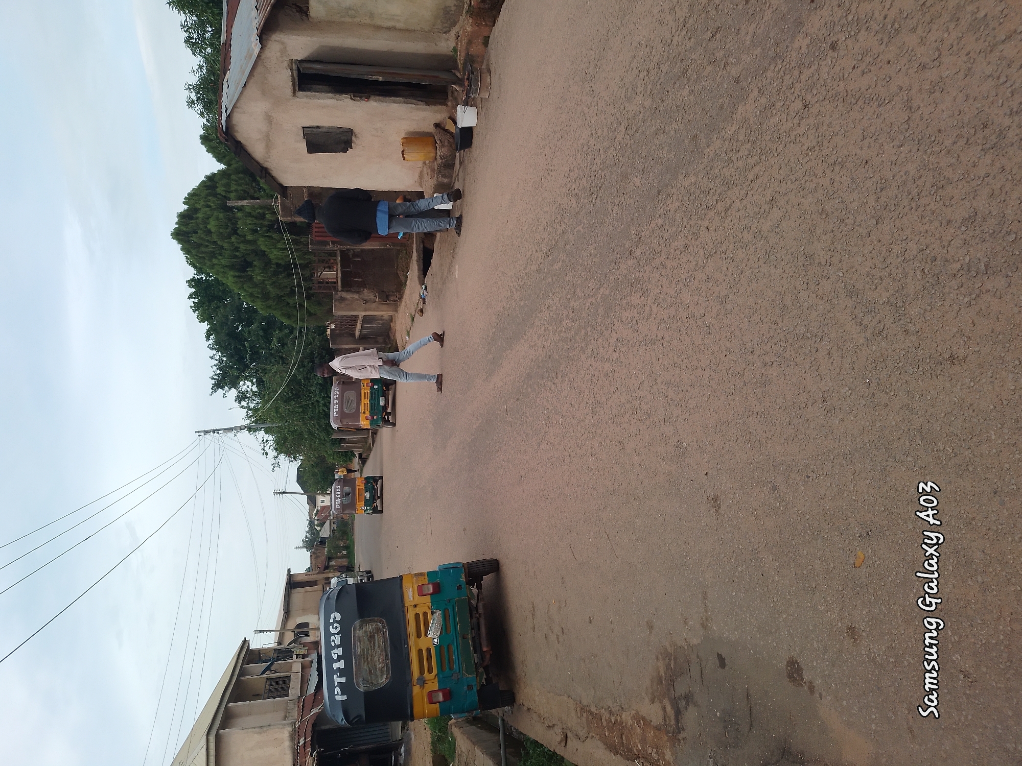 In Jos, Nigeria, a street scene unfolds under an overcast sky. A yellow and green auto-rickshaw, marked "PT-11265," is parked on the left side of the asphalt road. Further down the street, two other auto-rickshaws are visible, one with the marking "PTA 6021". Two men are walking on the right side of the road. One, wearing a light-colored shirt and blue jeans, walks away from the camera. The second man, in a dark jacket and blue jeans, walks towards the camera. To the right of the road, a small, dilapidated building with a corrugated metal roof stands. In front of the building are several objects, including a yellow container, two buckets, and what appears to be a small grill with burning coals. The environment appears to be a residential area within the city of Jos.