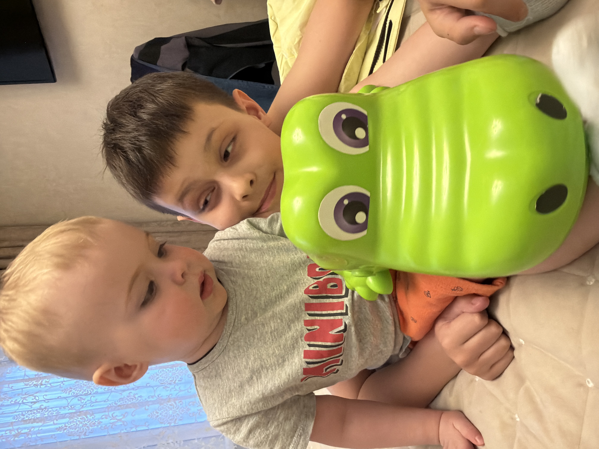 The image features two young boys interacting indoors, likely in Votkinsk, Russia. An older boy, with short brown hair and dark eyes, lies partially obscured by a large, bright green toy. He looks with a gentle, affectionate expression towards a younger child. The younger child, a baby with blond hair, is positioned in the foreground, wearing a grey t-shirt with red and black text that clearly spells out "WINNIE". He appears to be looking down or slightly to the side, his attention drawn to something outside the frame or the toy.
The prominent green toy, possibly a crocodile or dinosaur, has large purple and white eyes and is situated between the two children, indicating a play scenario. The older boy is wearing a yellow top, while the baby has orange shorts visible beneath his shirt. Both children are resting on a light-colored, soft surface, possibly a bed or play mat. In the background, a blue patterned curtain or blinds are visible to the left, and a dark, indistinct object can be seen in the upper left corner. The photo is taken from an overhead perspective, capturing a tender moment between the siblings.