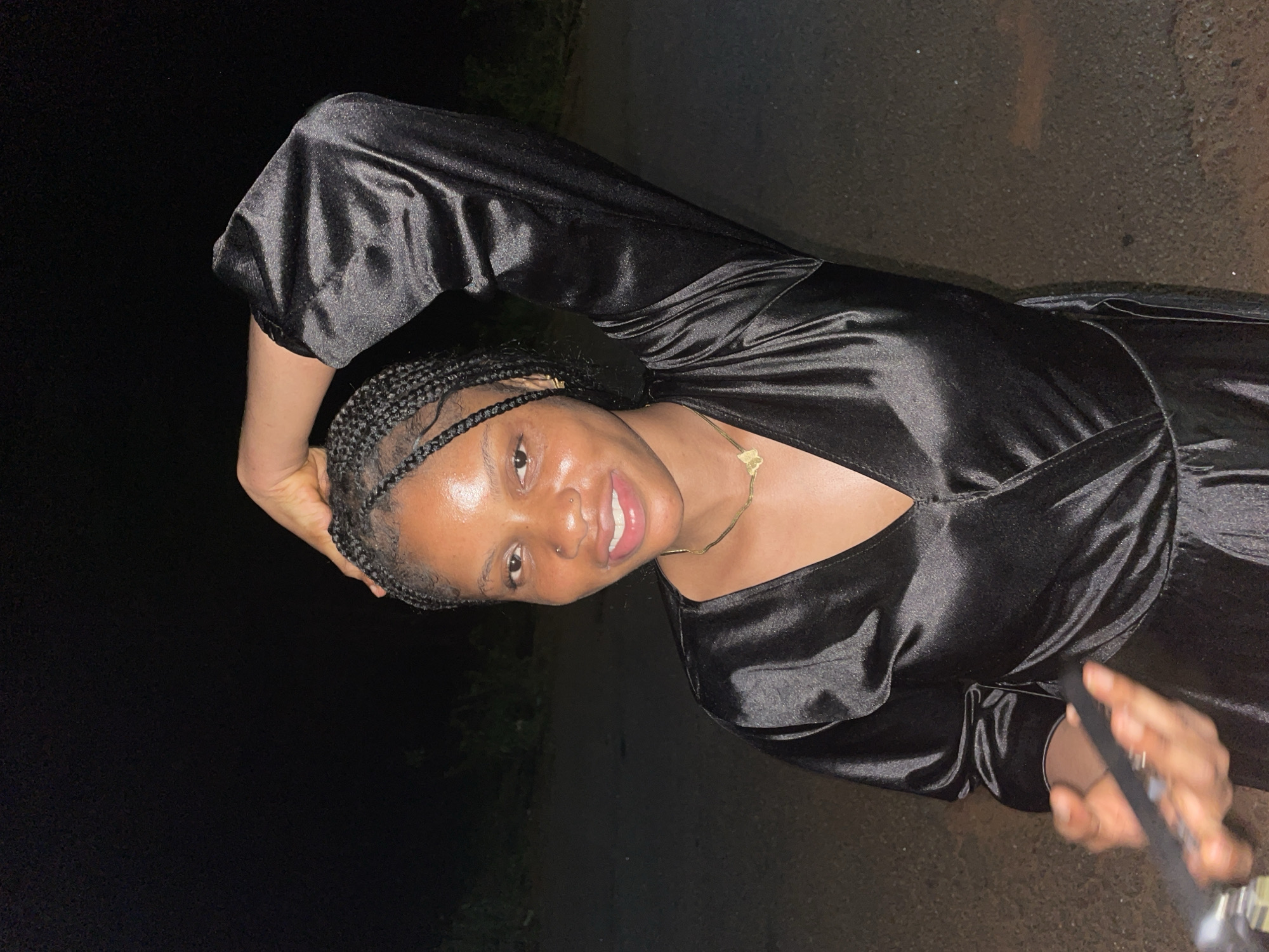 The image features a young woman with dark skin and braided hair, smiling broadly as she looks directly at the camera. She is wearing a shiny black satin-like dress or top with long, voluminous sleeves and a V-neckline. A delicate gold-colored chain necklace with a small pendant is visible around her neck, and she has a small piercing on her left nostril. Her left hand is raised, with fingers resting on the back of her head, while her right hand, partially visible in the bottom right, appears to be holding a camera or selfie stick.
The setting is outdoors at night, indicated by the predominantly dark background. The woman is well-lit, likely by a flash, which highlights her features and the glossy texture of her attire, contrasting sharply with the unlit surroundings. A dark, granular surface, possibly asphalt or concrete, is visible on the ground. Her expression conveys happiness and cheerfulness. No text is visible. The scene is located in Ogbe, Nigeria.