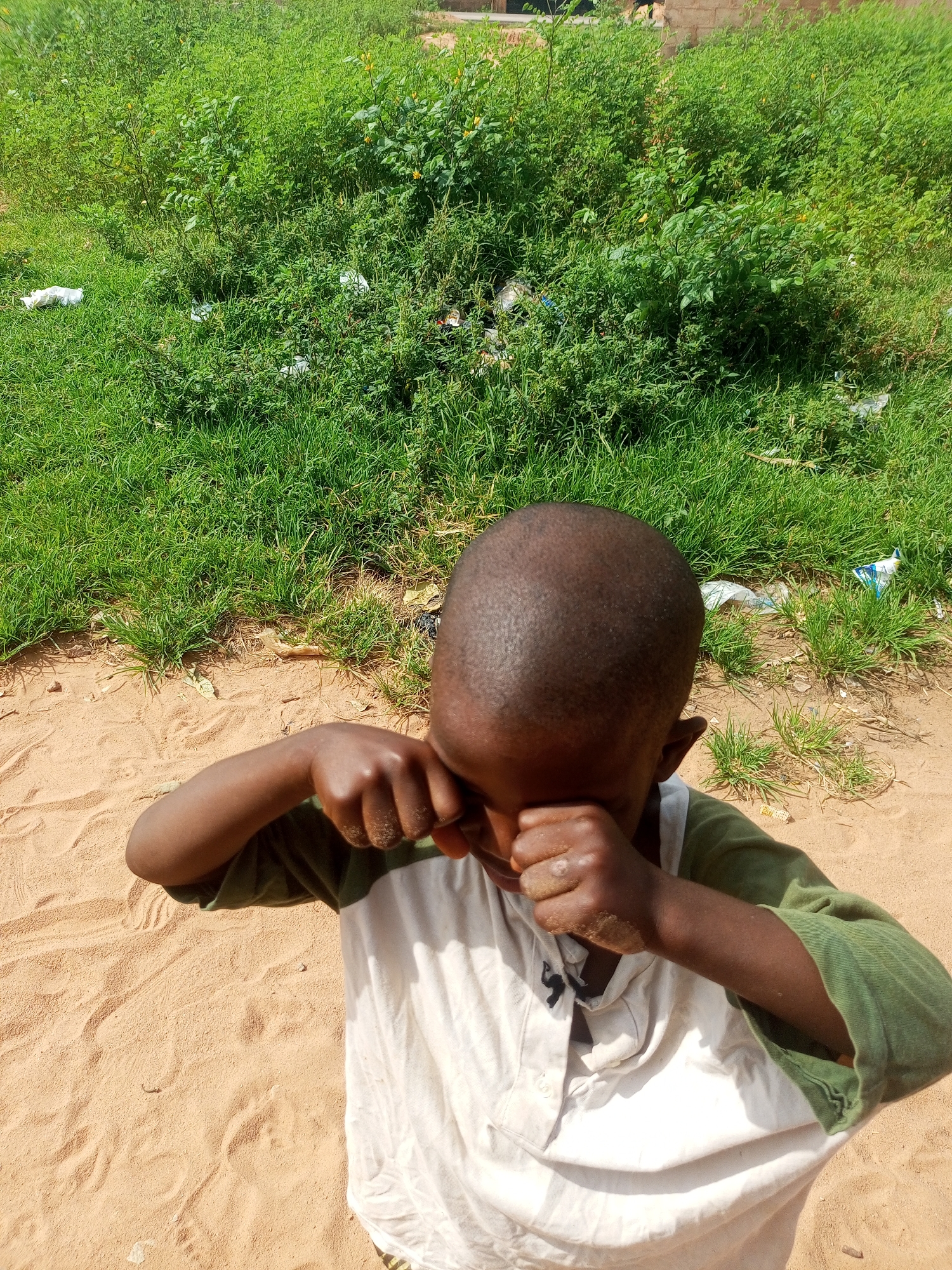 A young child, with dark skin and a shaved head, is kneeling on sandy ground in front of overgrown green vegetation. The child is wearing a white shirt with a green collar and sleeves, and is covering their face with their hands. The background consists of dense green plants, some of which appear to be weeds, with some scattered white debris visible among them. The ground is primarily composed of light brown sand, with some footprints and indentations visible. The lighting suggests it is daytime, with shadows indicating the sun is present. The scene appears to be in an outdoor, undeveloped area, possibly a vacant lot or the outskirts of a village.