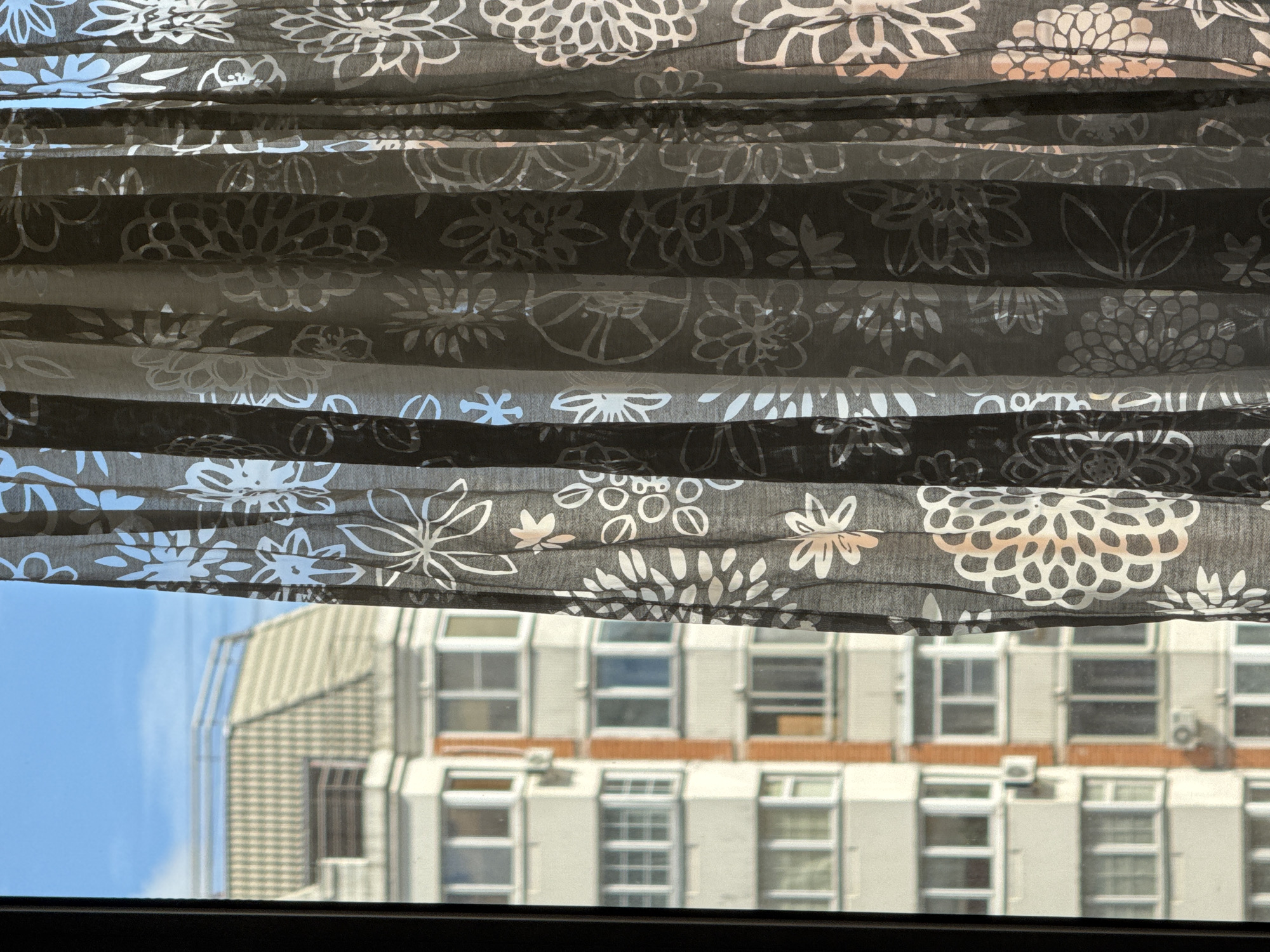 The image shows a view from a window in Moscow, Russia. In the foreground, a sheer dark gray curtain with a white floral pattern is hanging. The curtain is partially translucent, allowing light to pass through and illuminating the floral designs. In the background, a modern apartment building is visible. The building has multiple floors with numerous windows, many of which appear to be balconies. The exterior of the building is a light color, possibly white or light gray, with some sections showing reddish-brown brickwork. The sky above is blue and clear, with some faint clouds visible. The scene suggests a daytime view, likely from an apartment or office building.