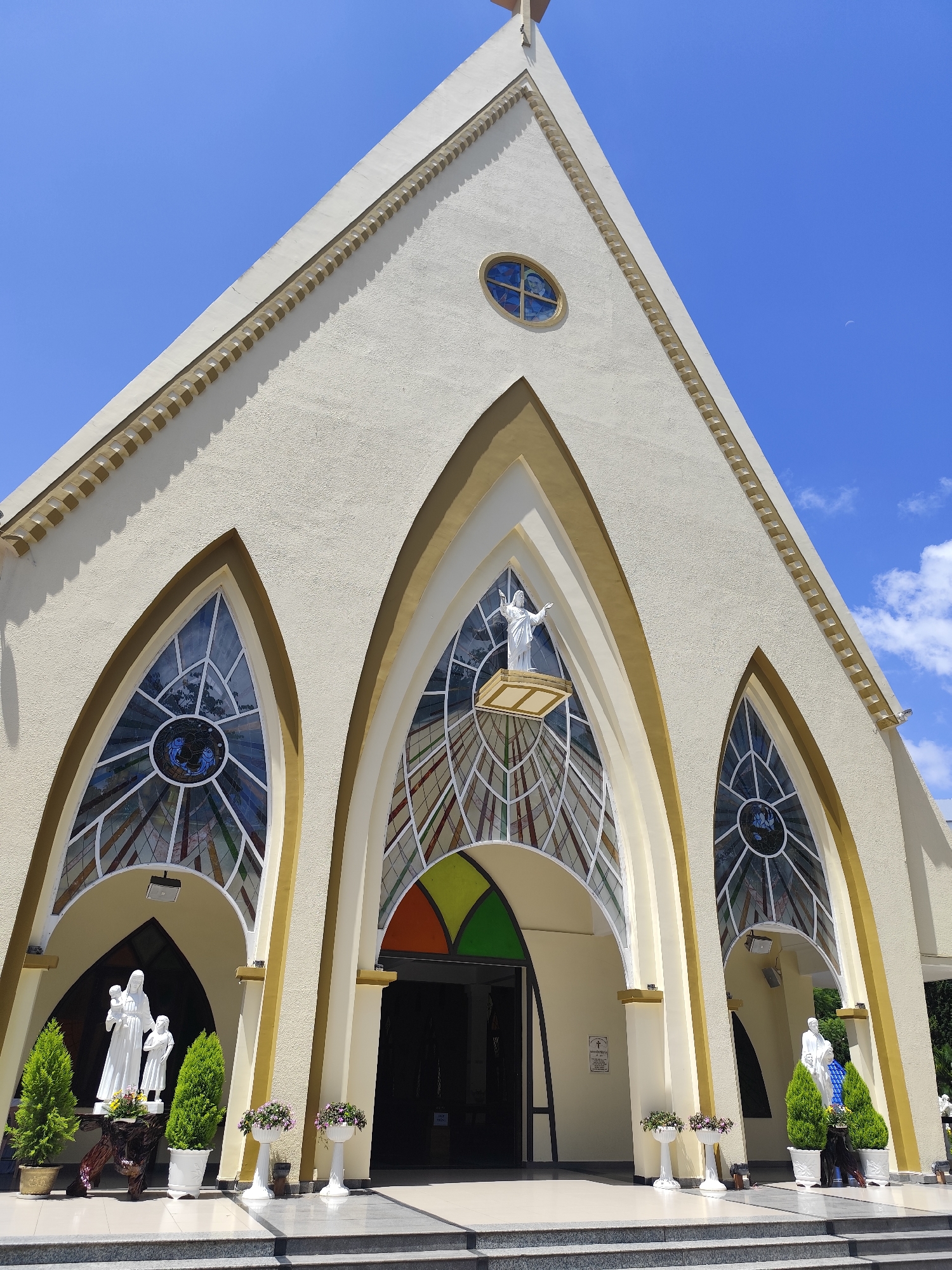 The image depicts the facade of a church, likely located in Da Lat, Vietnam, given the architectural style and prevalence of such religious structures in the area. The building is painted a light cream color with a pointed, triangular roofline. The entrance is grand, featuring a series of arched openings with decorative gold trim. Large, stained-glass windows are prominent features, with intricate patterns in blues, reds, and greens. Above the central entrance, a statue of Jesus Christ stands with arms outstretched. Flanking the entrance, on either side of the walkway, are white statues of religious figures, possibly Mary and child on the left. These statues are placed near potted evergreen trees and flower arrangements. The ground in front of the church is paved with tiles, leading up to a set of stone steps. The sky above is a vibrant blue with a few scattered white clouds. The overall impression is one of a well-maintained and architecturally significant religious building under a clear sky.