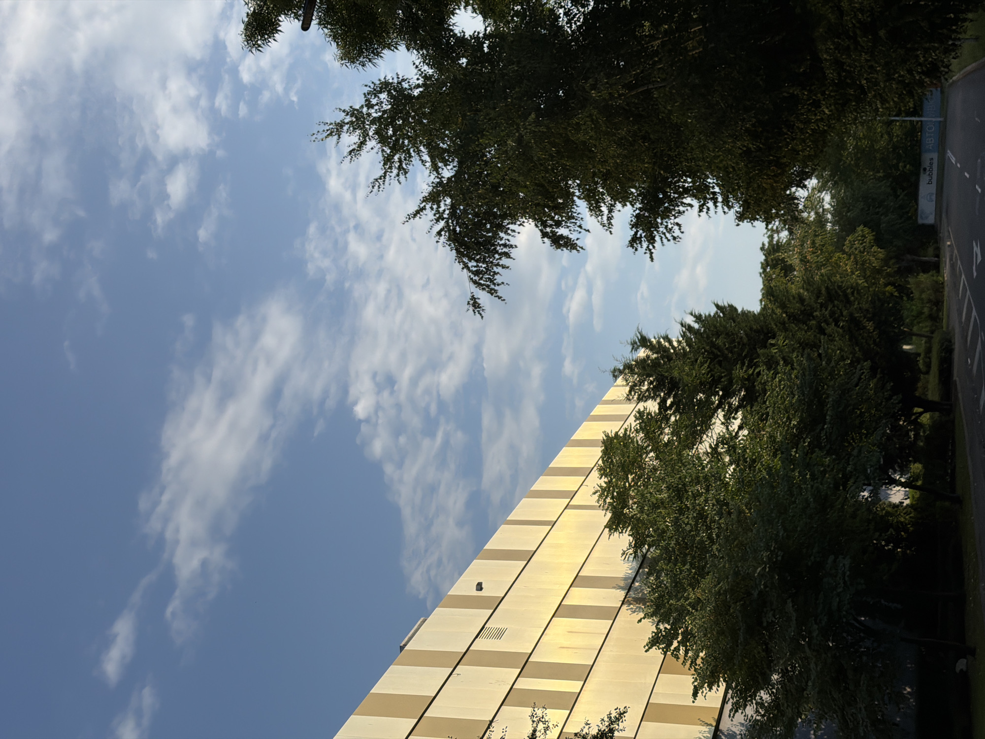 The image depicts a modern building with a golden facade, characterized by vertical stripes of varying shades of yellow and beige. The building is partially obscured by lush green trees in the foreground. A paved road leads away from the viewer into the distance, with visible lane markings. In the background, a sign reads "bubbles ABTO" in blue and white lettering. The sky above is a clear blue, dotted with wispy white clouds. This scene is located on a street in Burgas, Bulgaria.
