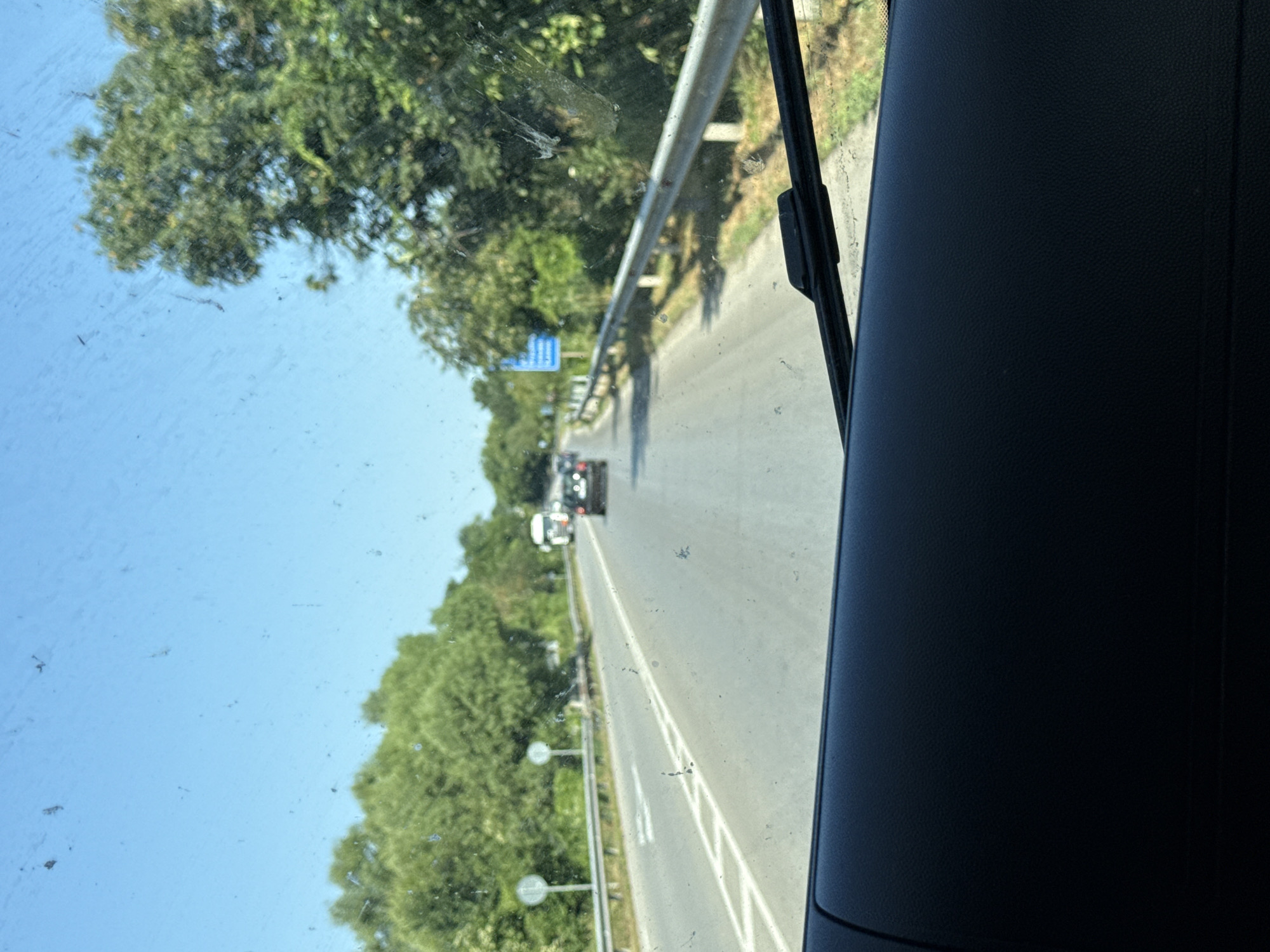 The image depicts a view through a car windshield on a clear, sunny day. The road ahead is paved with white lane markings. A dark-colored car is in front, and a white truck is further down the road. Trees line the sides of the road, and a metal guardrail runs alongside it. A blue sign is visible in the distance. The foreground of the image shows the dark dashboard of the vehicle.
While the image itself does not explicitly confirm the location as Osenets, Bulgaria, the presence of road signs and guardrails suggests a roadway in a populated area. Further information would be needed to definitively pinpoint the location within Osenets.