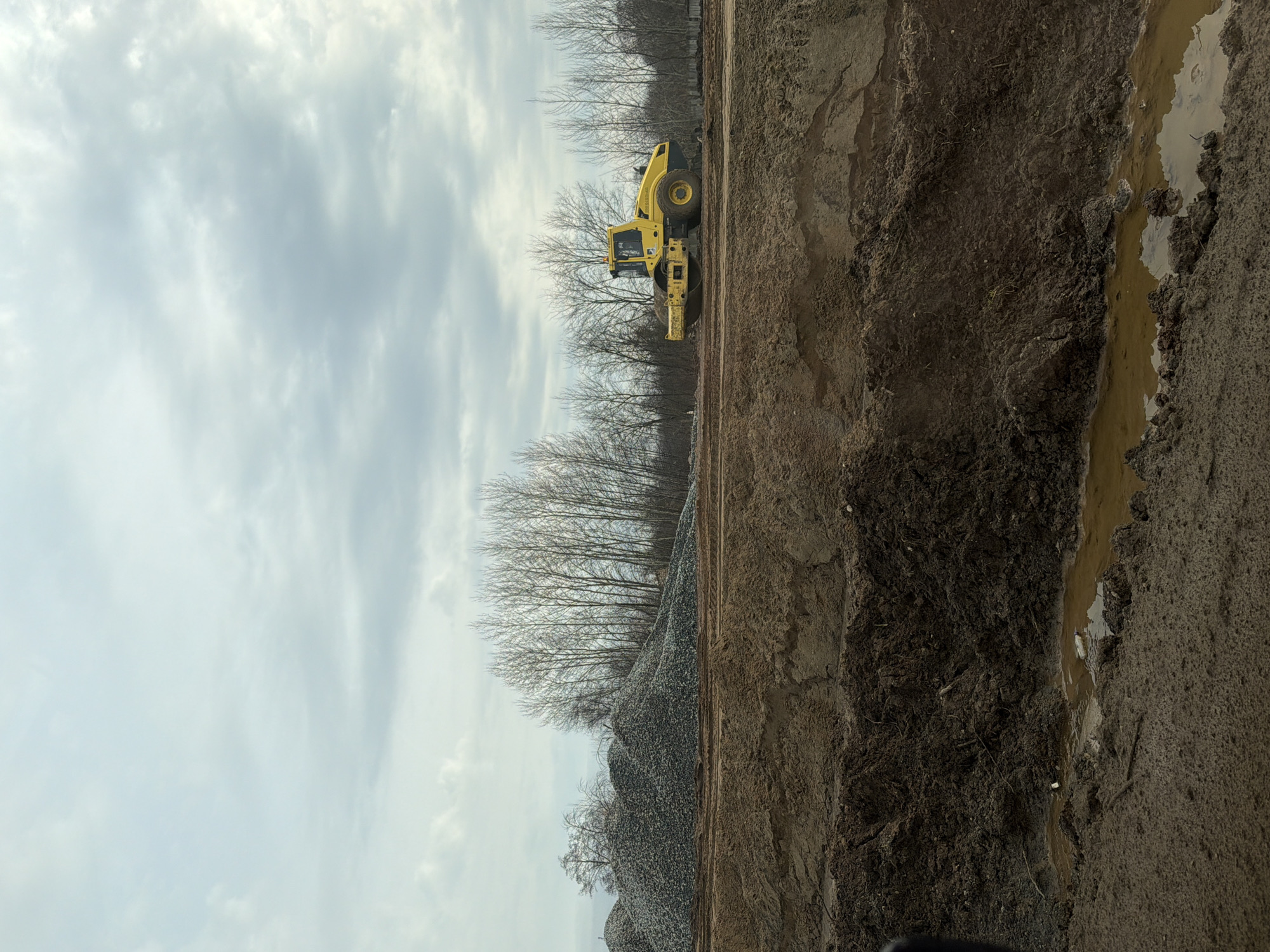 The image shows a construction site in Zav'yalovo, Russia. A yellow road roller is positioned next to a pile of gravel, with bare trees in the background. The foreground contains a muddy, dug-out area with standing water. The sky is overcast.