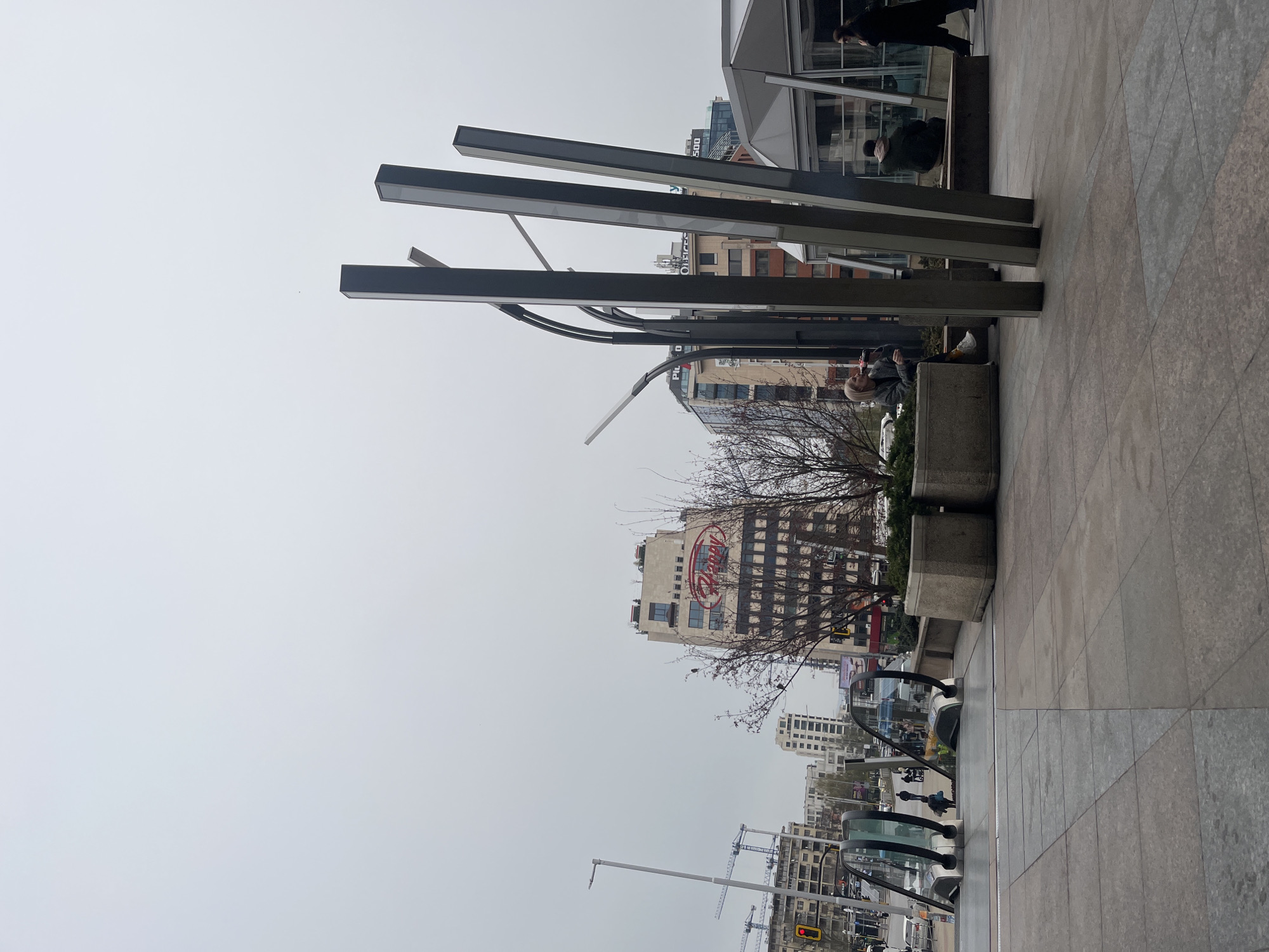 The image depicts a street scene in Sofia, Bulgaria. The foreground features a pedestrian walkway paved with gray rectangular tiles, lined with low concrete planters containing trees and greenery. Behind the planters, a series of tall, gray, rectangular metal structures rise vertically, creating a modern architectural element. Further back, a building with a prominent "Darby" sign is visible. People are sitting and standing around the planters and near the metal structures. Several buildings are situated beyond that, including apartment blocks and commercial structures, all set against a cloudy sky. In the lower part of the image, a road with vehicle traffic, streetlights, and construction cranes in the distance are visible.