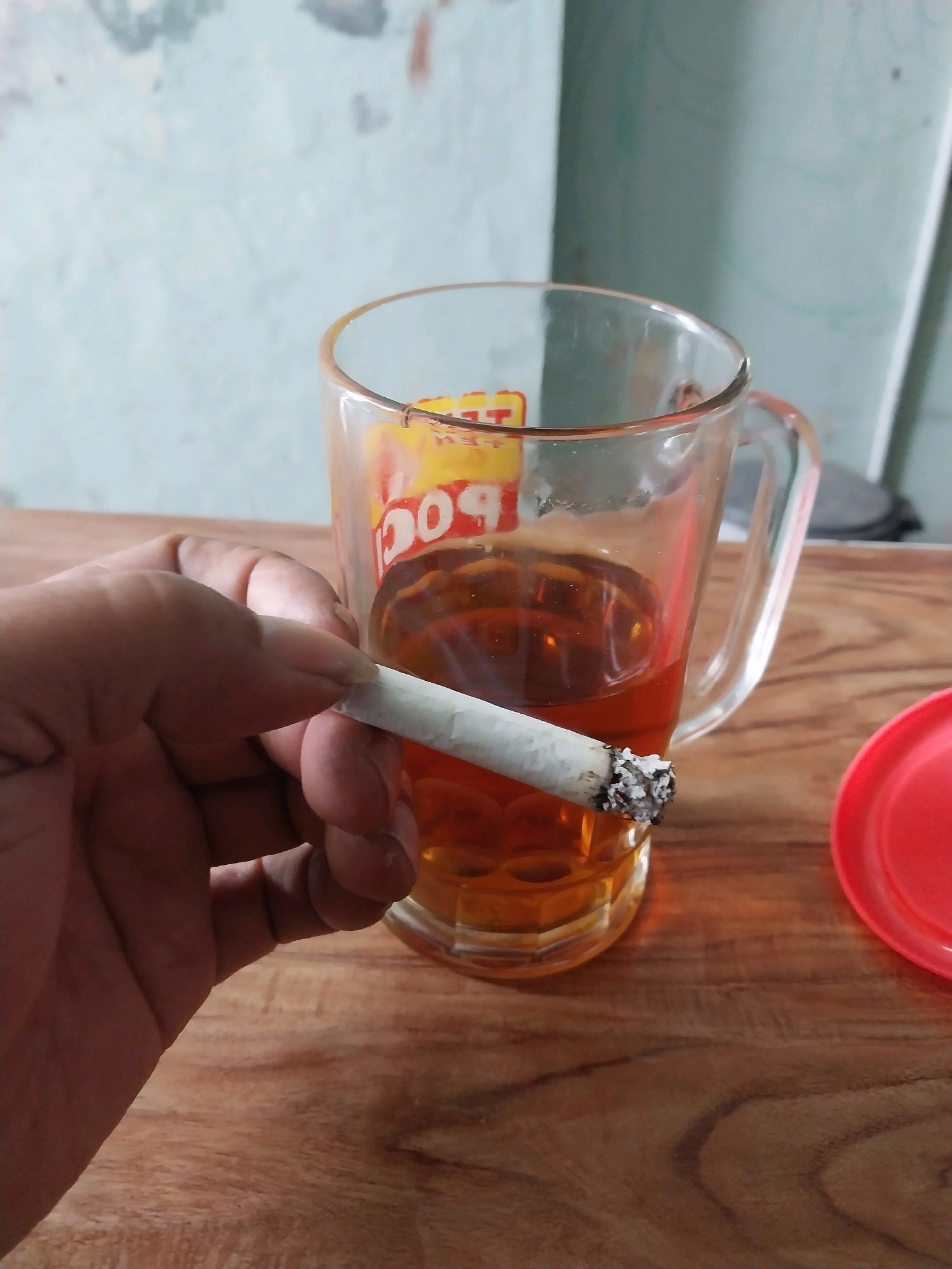 A close-up shot shows a person's hand holding a lit cigarette over a glass of amber-colored liquid, likely tea, on a wooden surface. The glass has a visible logo with red and yellow lettering. A red lid is partially visible to the right of the glass. The background is a light-colored wall with an abstract pattern. The scene appears to be indoors, possibly in a home or a simple establishment in Wates, Indonesia, suggesting a moment of relaxation or a break.
