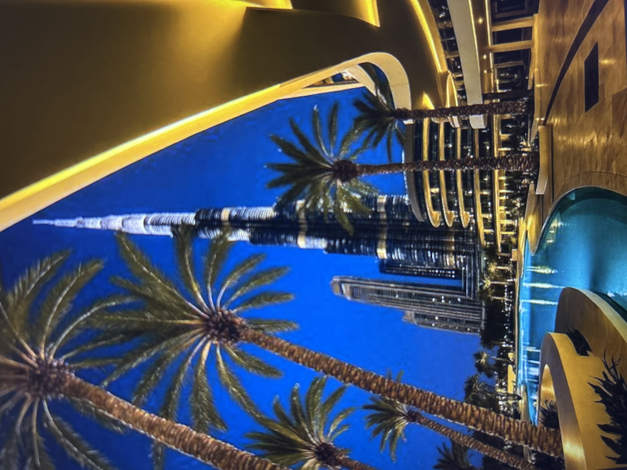 This image captures a vibrant night scene in Dubai, United Arab Emirates, featuring the iconic Burj Khalifa dominating the skyline. In the foreground, a winding, brightly illuminated swimming pool or water feature with glowing blue water is bordered by curvilinear, golden-hued architectural elements and lush palm trees. The pool area is paved with reflective surfaces, suggesting a resort or luxury residential complex.
Several mature palm trees with textured trunks and prominent fronds are scattered throughout the mid-ground and foreground, providing a tropical accent against the deep blue, twilight sky. To the right, a multi-story, circular modern building with numerous warmly lit windows and balconies is visible, part of the extensive complex.
In the background, other contemporary skyscrapers stand, contributing to the urban panorama. The Burj Khalifa, positioned centrally, is distinctively lit, highlighting its intricate architectural details and soaring spire against the dark sky. The overall environment is one of modern luxury and sophistication, characterized by sleek design, abundant lighting, and well-maintained landscaping. No individuals are visible in the scene.