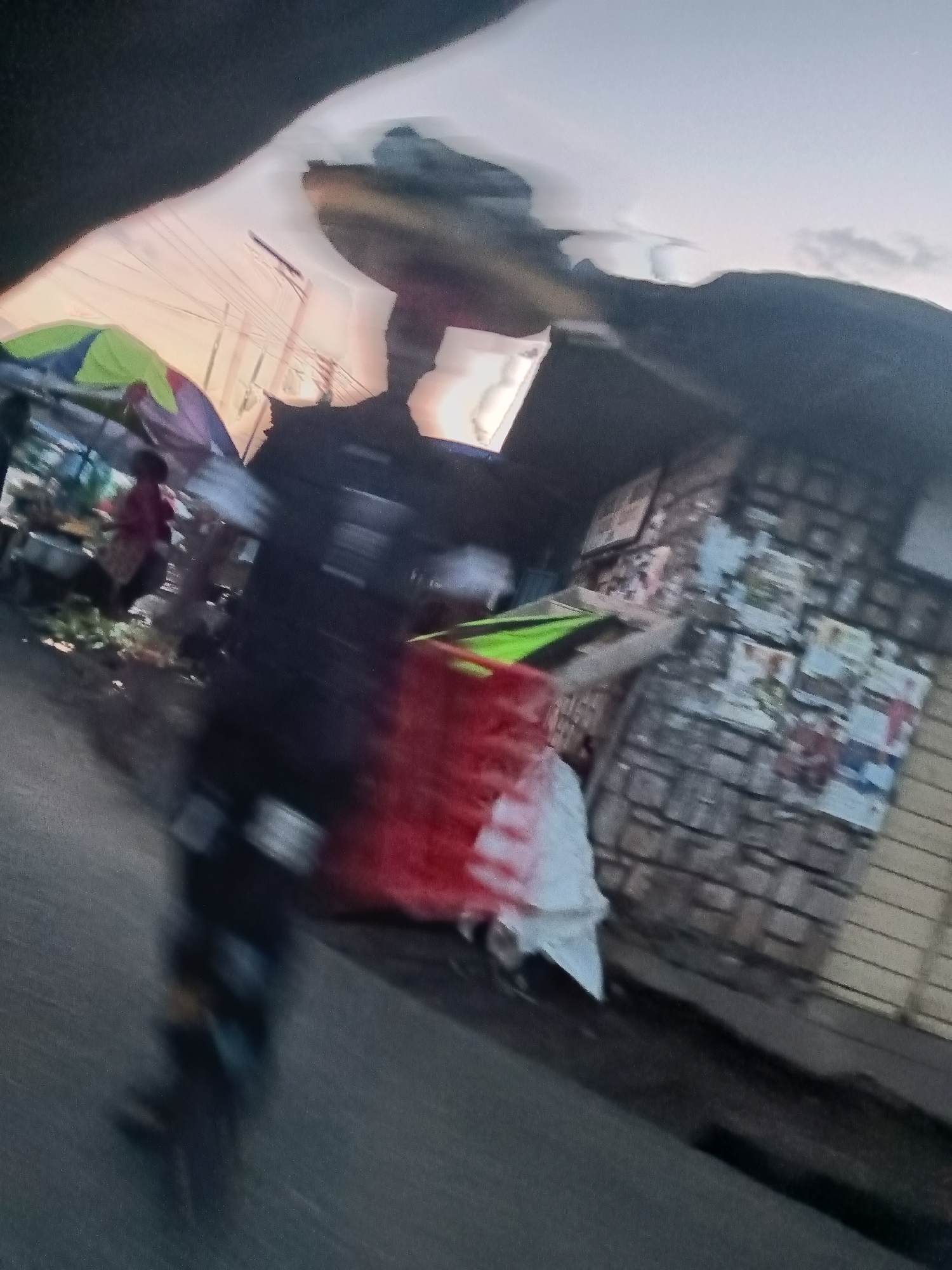 A blurred, low-angle, medium shot depicts a street scene at dusk in Jos, Nigeria. The foreground is dominated by a dark, blurry shape, possibly a vehicle or person on a motorcycle, with a bright red object visible behind it. To the left, a market stall with a green and yellow canopy is partially visible, with a person in a red garment standing behind it. In the background, a brick wall is covered with posters and flyers, with a corrugated metal structure to its right. The sky above is a pale orange and grey, suggesting sunset. The overall impression is one of a busy, urban environment.