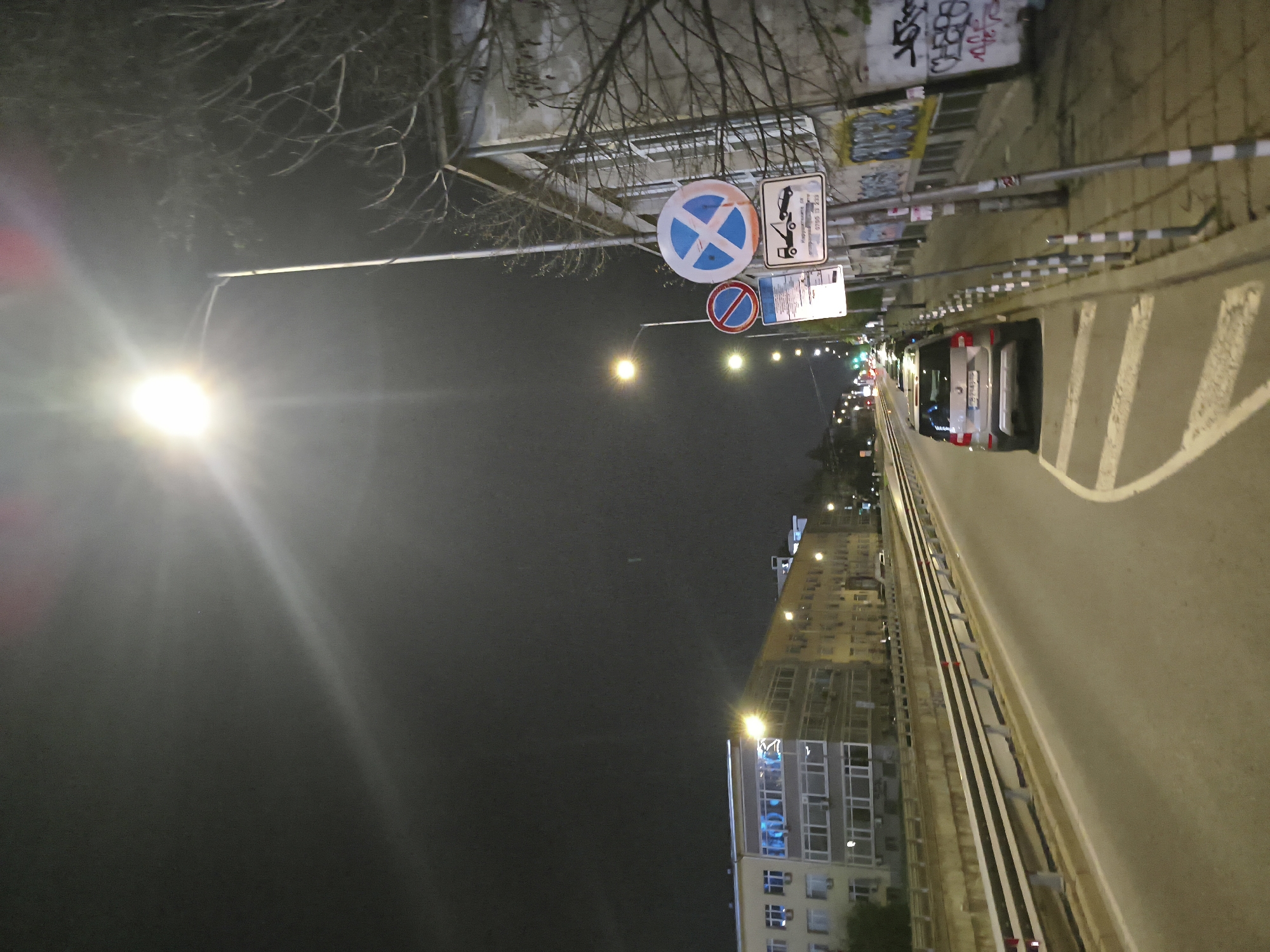 The image shows a street scene in Sofia, Bulgaria, at night. A white car is driving on the right side of the road. Traffic signs indicating "no stopping" and "no parking" are visible. Graffiti is present on the wall beside the street. A tall building with lighted windows is seen in the background. The street is illuminated by streetlights. Bare tree branches are in the upper left corner of the image.