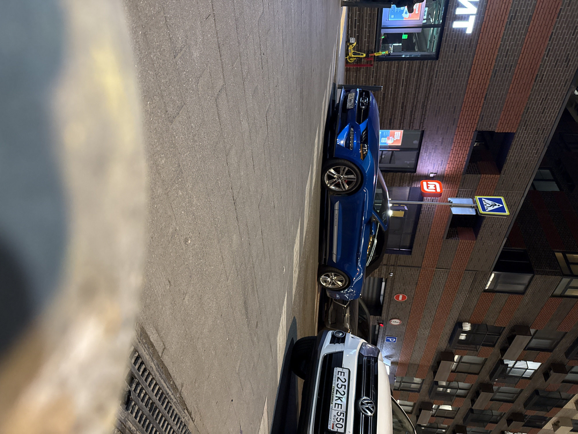 The image shows a street scene in Khimki, Russia at night. A blue Ford Mustang is parked on the street, alongside a white Volkswagen vehicle. The Volkswagen has a license plate that reads "E252KE 550 RUS." The buildings nearby have brick facades with dark window recesses. A pedestrian crossing sign is visible, and there is signage in Cyrillic script. Street lighting illuminates the area. A metal grate is visible in the foreground. No people are visible.