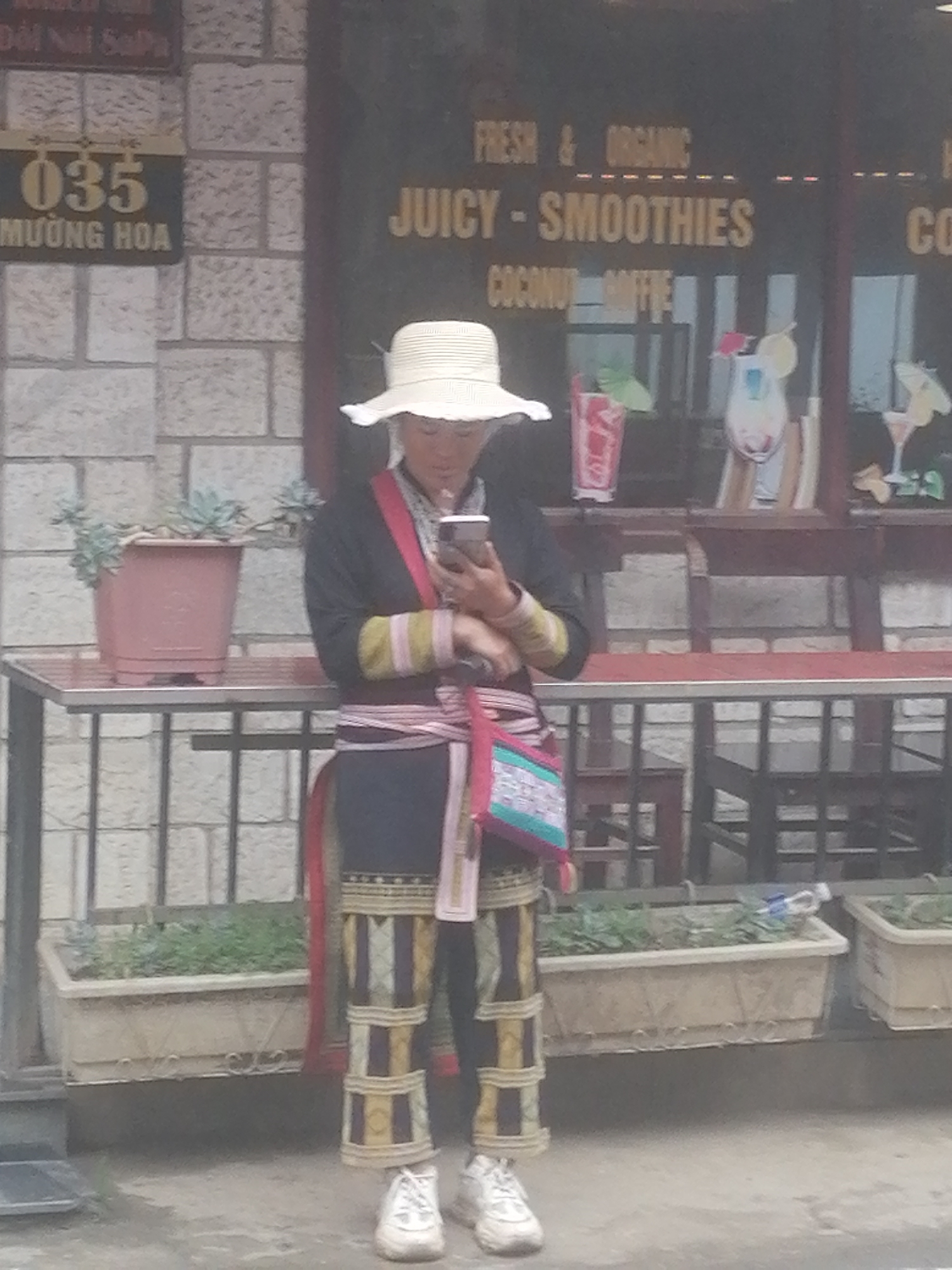 This full-shot image features a woman in traditional attire, engrossed in her smartphone, standing outside a beverage shop in Sa Pa Ward, Vietnam. The woman, positioned centrally, wears a wide-brimmed light-colored hat, a dark long-sleeved top with colorful striped cuffs in green, yellow, and pink, and dark pants patterned with gold, dark blue, and white geometric designs on the lower legs. A multi-colored sash, primarily pink and white, is wrapped around her waist, and she carries a vibrant patterned shoulder bag with pink, light blue, and white elements. White sneakers complete her outfit.
She holds a smartphone with both hands, looking down intently at its screen. To her left, against a light-colored brick wall, is a brown pot containing succulents, placed on a metal table. A sign on the wall displays "035 MƯỜNG HOA" in yellow text, with fainter text "Dồi Núi SaPa" above it.
The background on the right reveals the dark facade of a shop, with prominent yellow text advertising "FRESH & ORGANIC JUICY - SMOOTHIES COCONUT COFFEE". Decorative illustrations of colorful drinks in glasses, some with small umbrellas, are visible through the window. Inside the shop, glimpses of tables and chairs can be seen. Along the base of the shop, several rectangular planters contain green foliage. The ground is a light-colored paved surface. The scene captures a blend of traditional culture with contemporary life in a daytime setting.